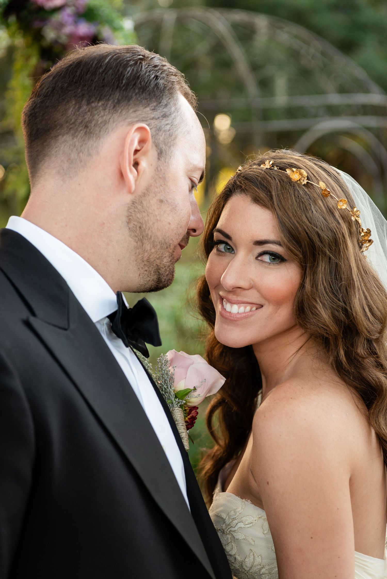 Smiling bride wearing gold leaf crown and lace gown with groom in black tuxedo during outdoor wedding portraits.