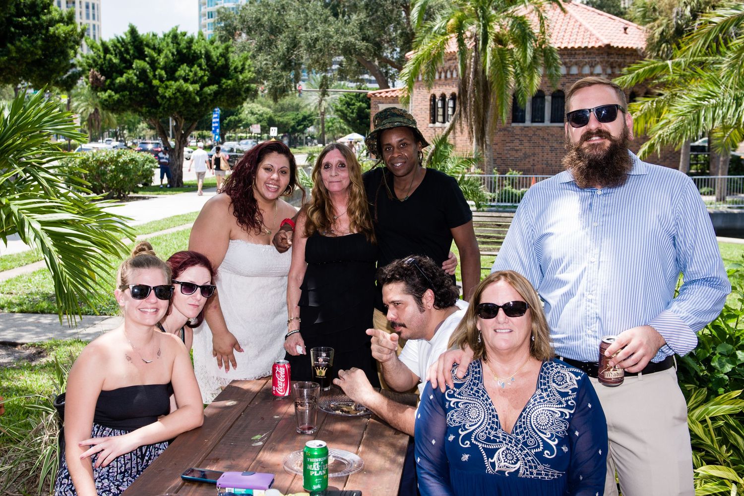 Group of friends gathered around a picnic table outdoors at a sunny park event, smiling and socializing together.