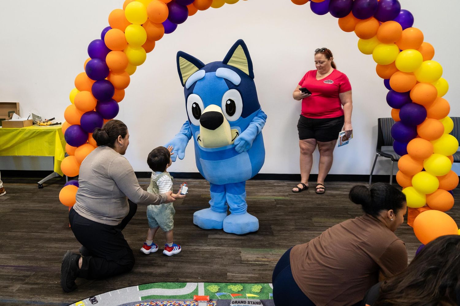 A toddler meets a Bluey mascot character under a colorful balloon arch at a birthday party.