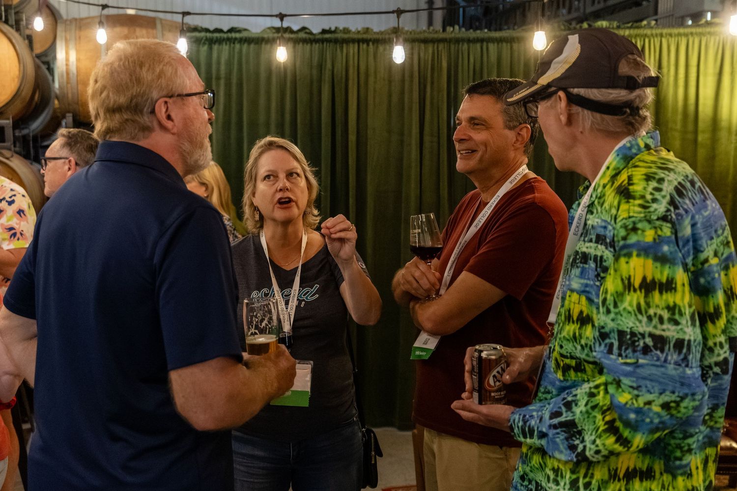 Group of adults socializing at an indoor craft beer festival event with string lights and barrels in background.