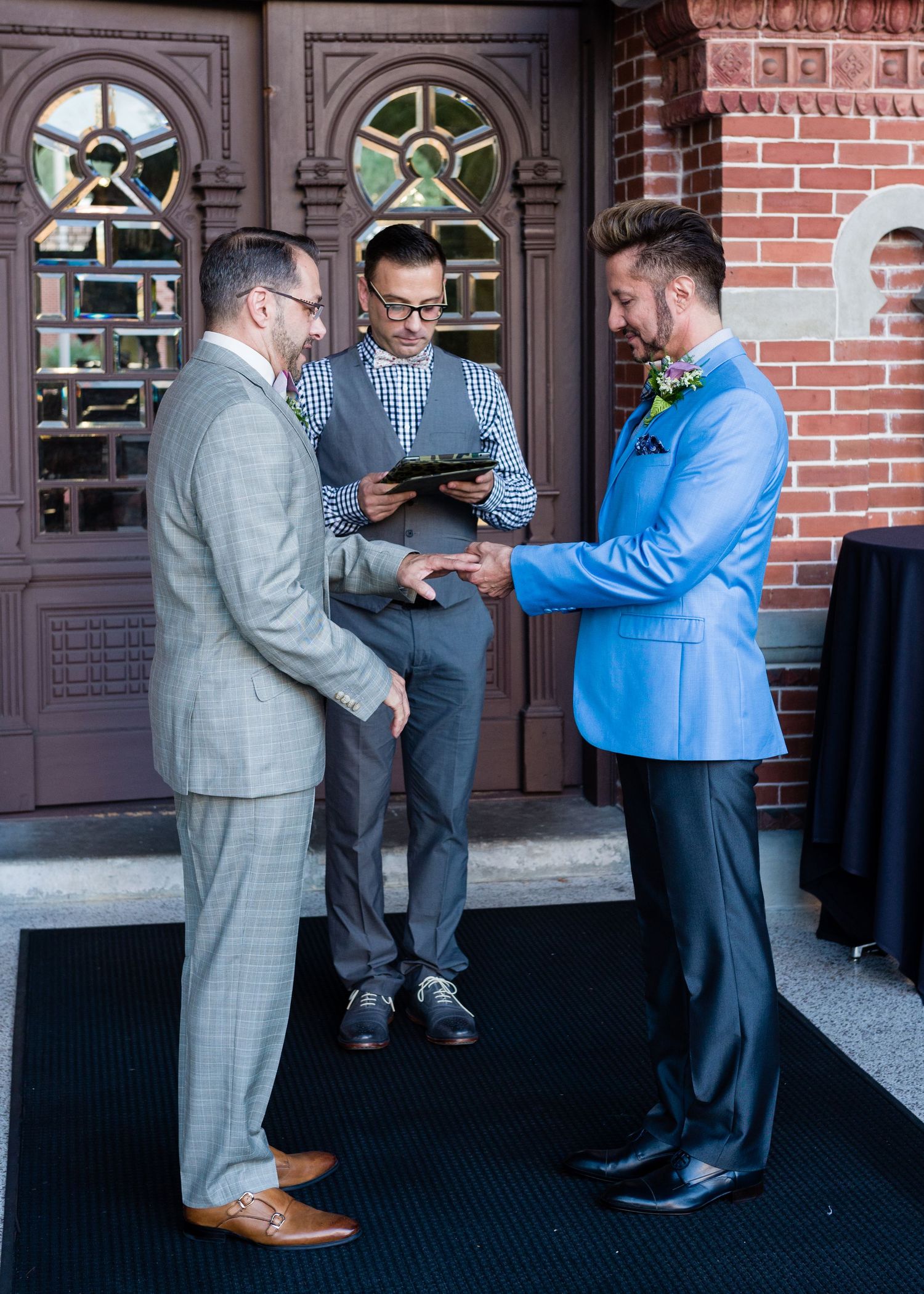 Two grooms exchange rings during outdoor same-sex wedding ceremony.