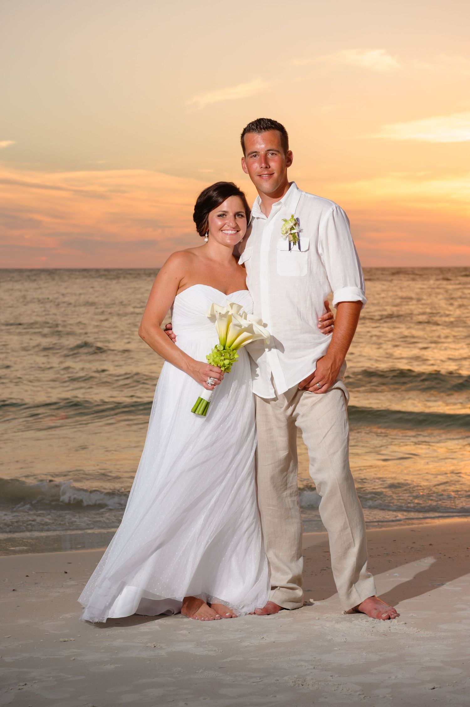 Smiling bride and groom posing barefoot on a sandy beach during a vibrant sunset.