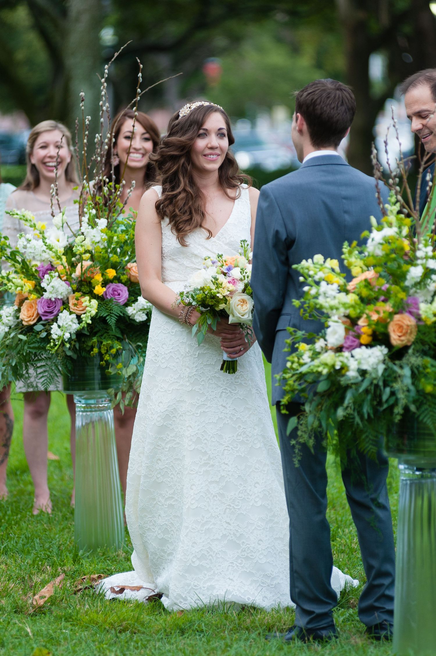 Smiling bride in white lace gown stands at altar surrounded by lush floral arrangements during outdoor ceremony Museum of Fine Arts, St. Pe.