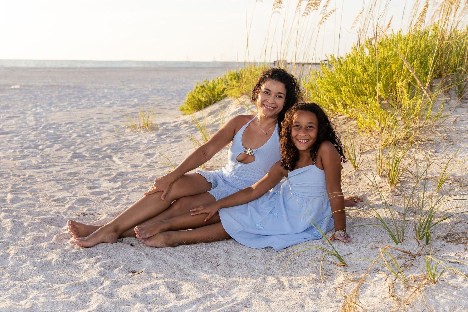 Two young women in white dresses sit together on sandy beach near grass dunes, smiling warmly at camera.