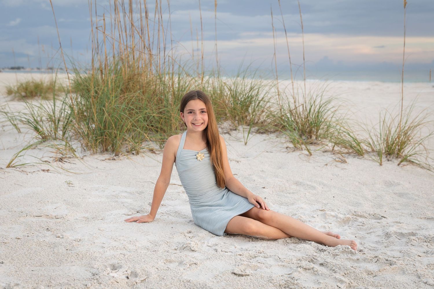A young girl in a light blue dress sits on white sand beach with sea oats and dunes in the background.
