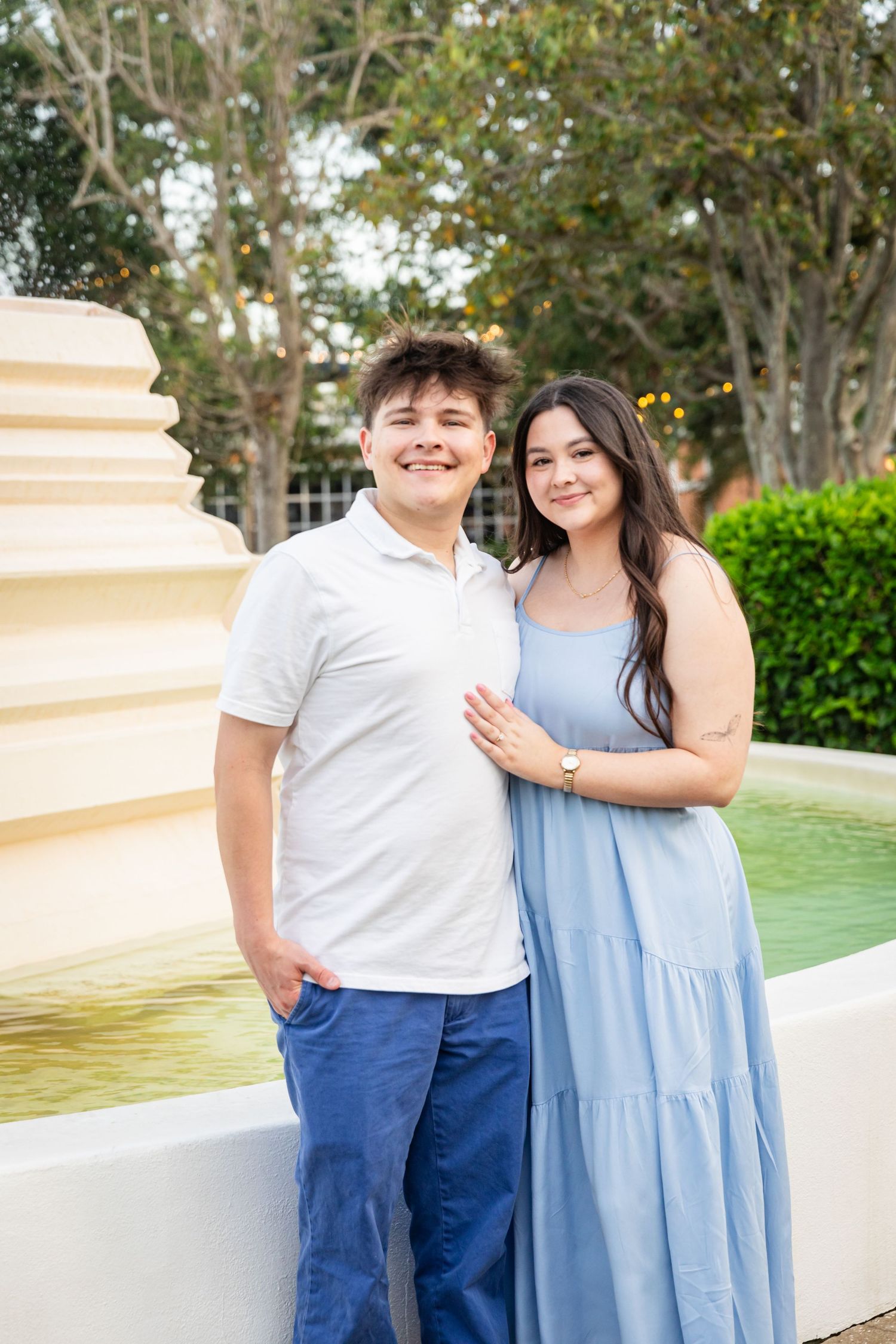 Smiling young couple posing by a decorative fountain outdoors, man in white polo and woman in blue maxi dress.