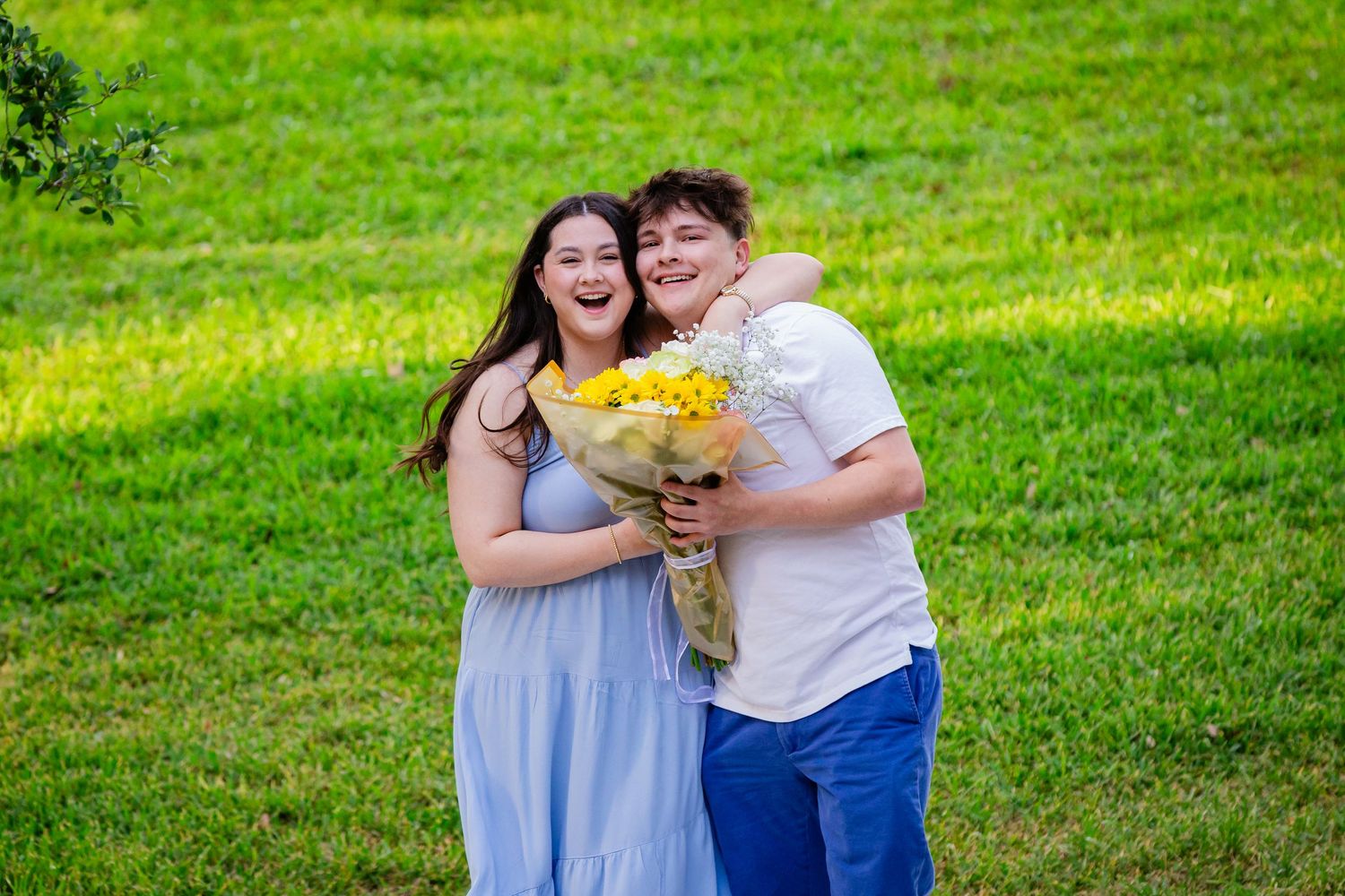Two smiling teens pose outdoors on green grass, sharing a bouquet of yellow flowers in a joyful embrace.