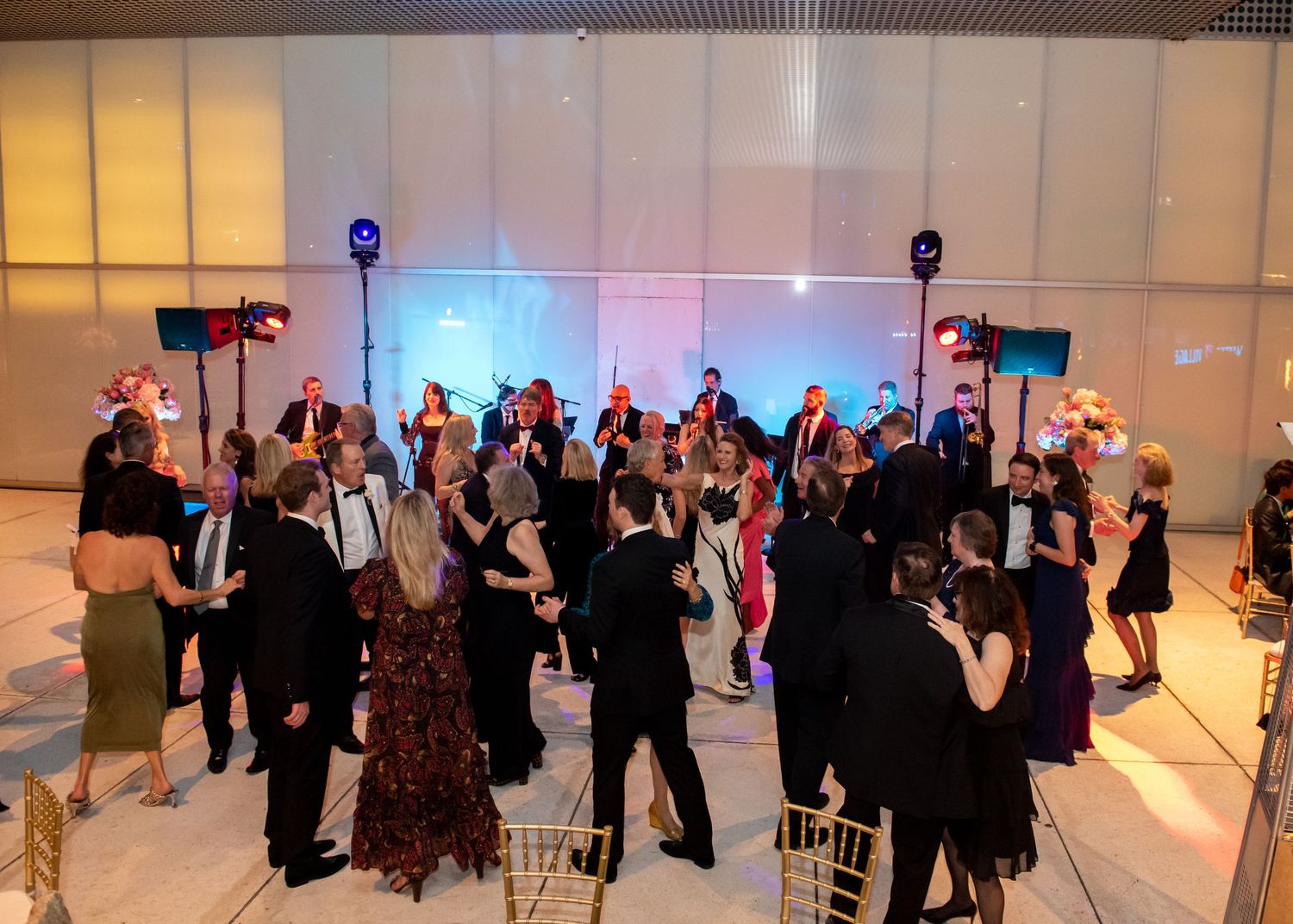 Elegantly dressed guests dancing and socializing at a formal gala event in a modern venue with stage lighting.
