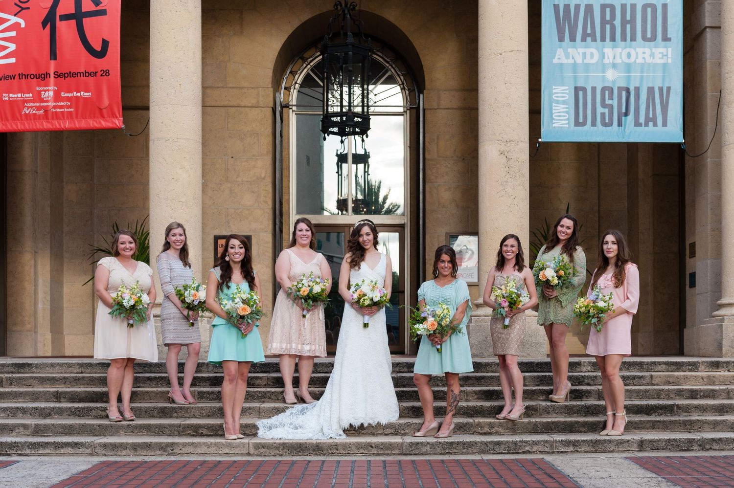 Bride with bridesmaids in mint and blush dresses holding bouquets outside a museum entrance.
