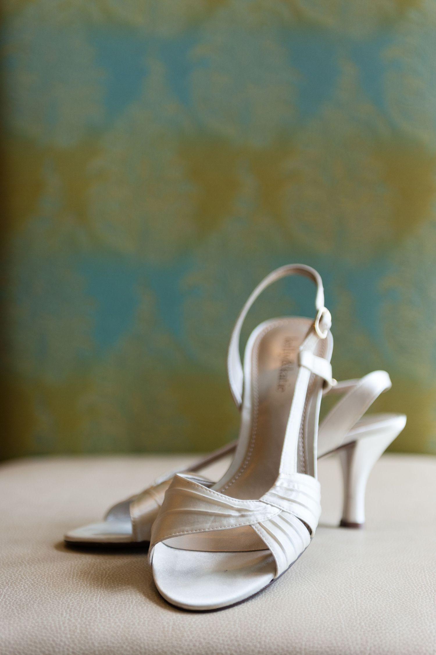 Elegant white satin slingback wedding heels displayed on a cream surface with decorative wallpaper background.