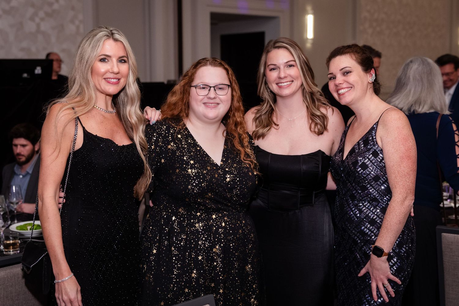 Four women in elegant black dresses posing together at a formal gala event.