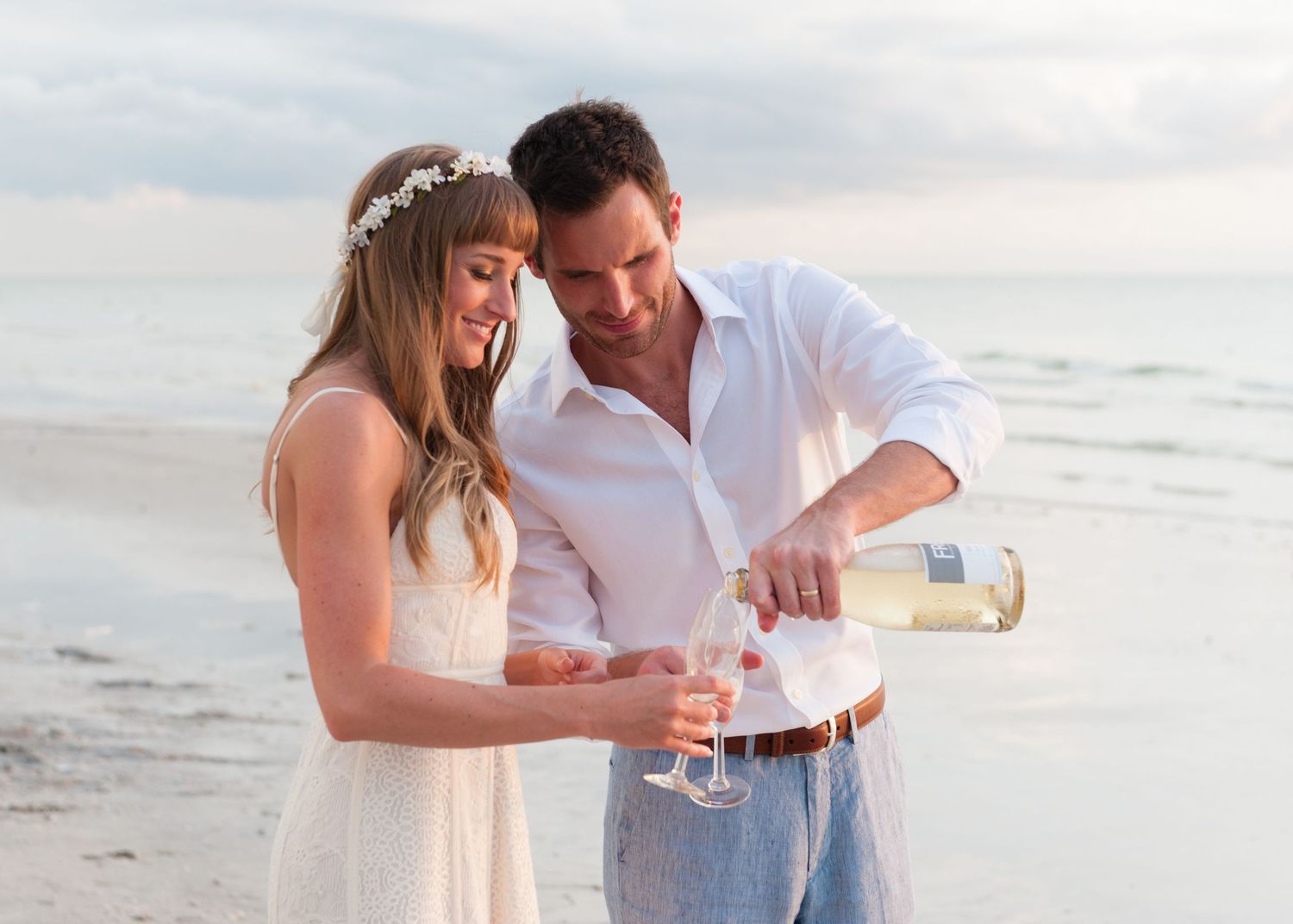 Couple in white attire pours champagne together on a beach during a romantic seaside wedding celebration.