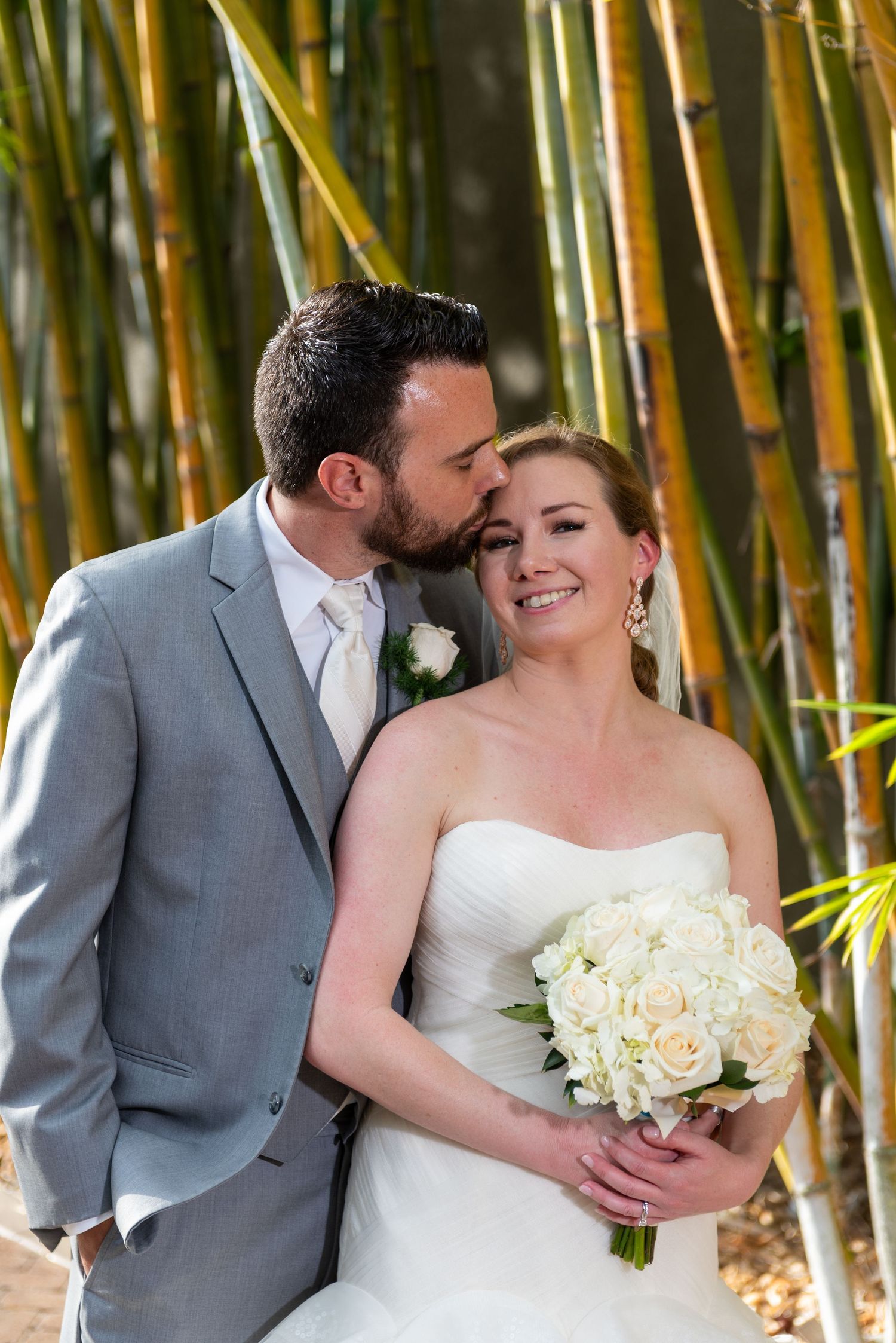 Groom in gray suit kisses bride's cheek as she smiles holding white rose bouquet among bamboo trees.