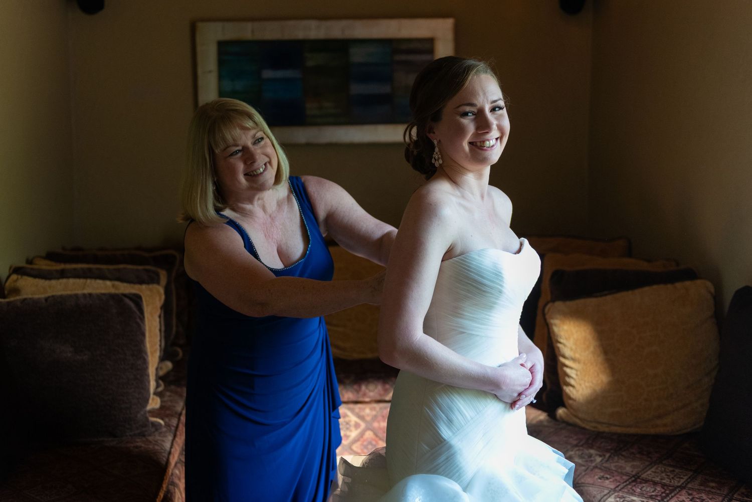 A woman in a blue dress helps a smiling bride in a white strapless gown get ready in a warmly lit room.