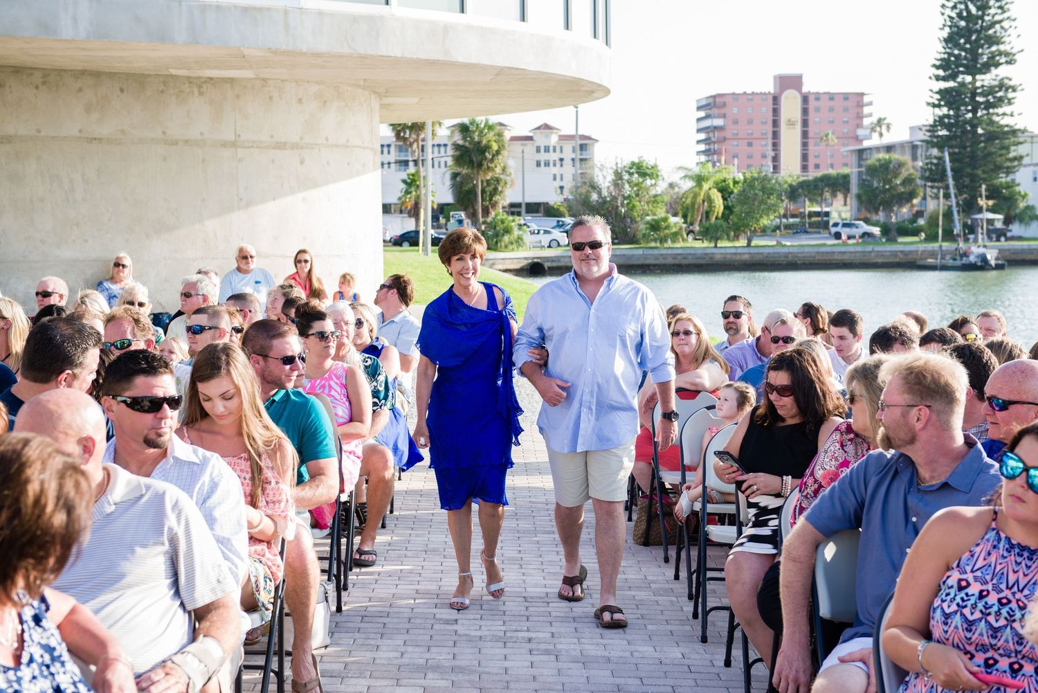 Couple walks down outdoor wedding aisle by waterfront, guests seated in chairs under sunny blue sky.