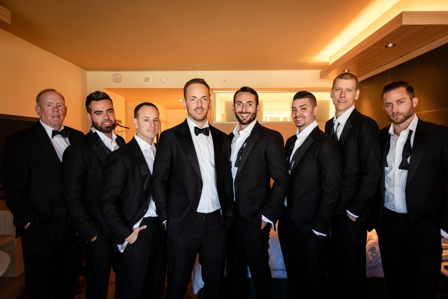 Group of eight groomsmen wearing black tuxedos and bow ties posing together indoors for a wedding photo.