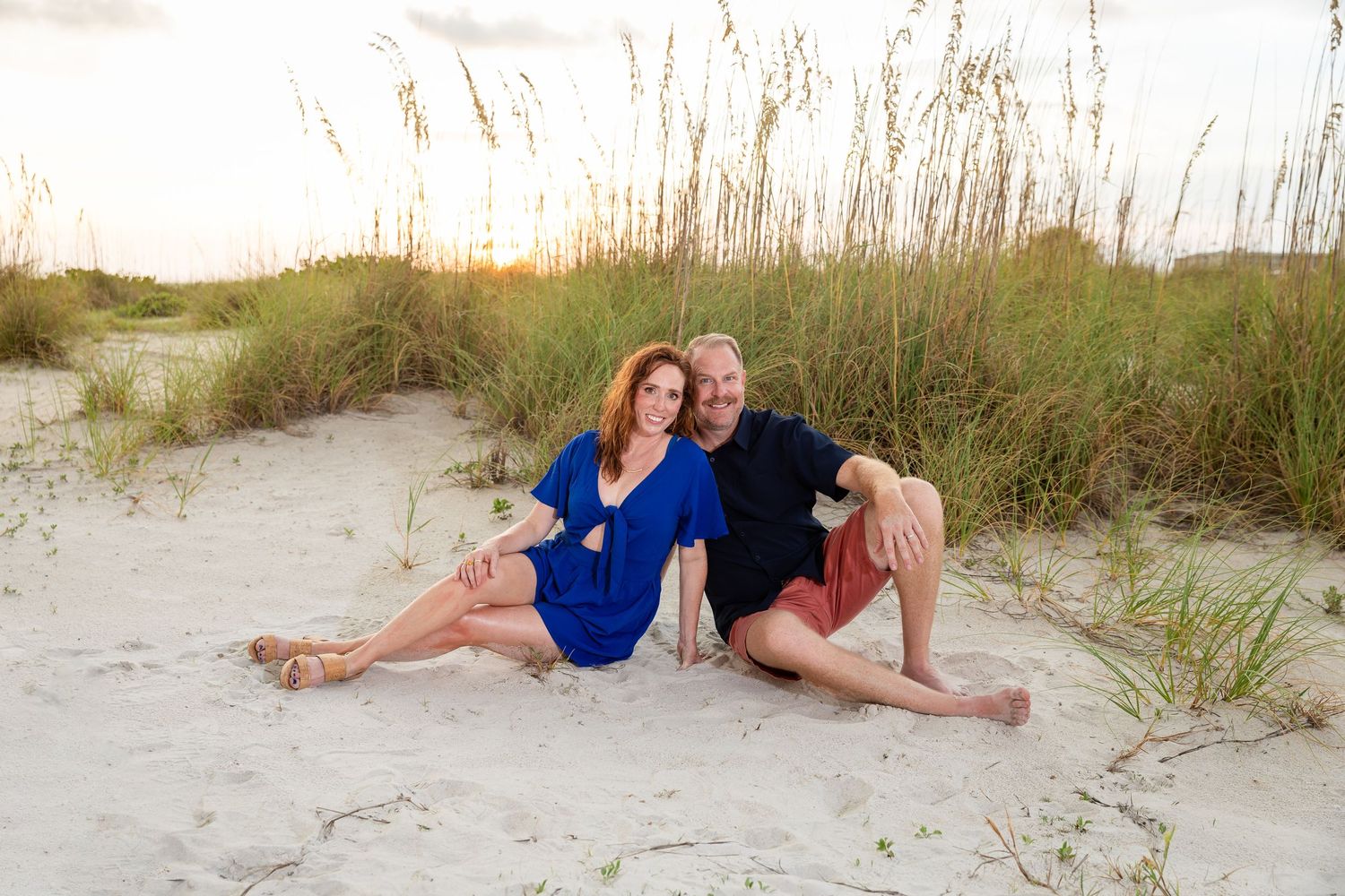 Couple sitting together on sandy beach dunes with sea oats, woman in blue dress, man in black shirt and red shorts.