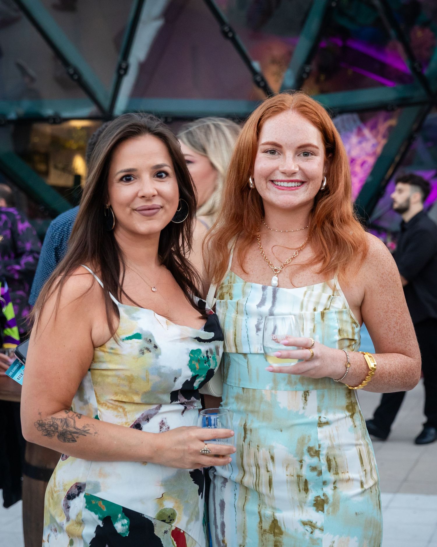 Two smiling women in floral and pastel dresses holding drinks at an outdoor evening event.