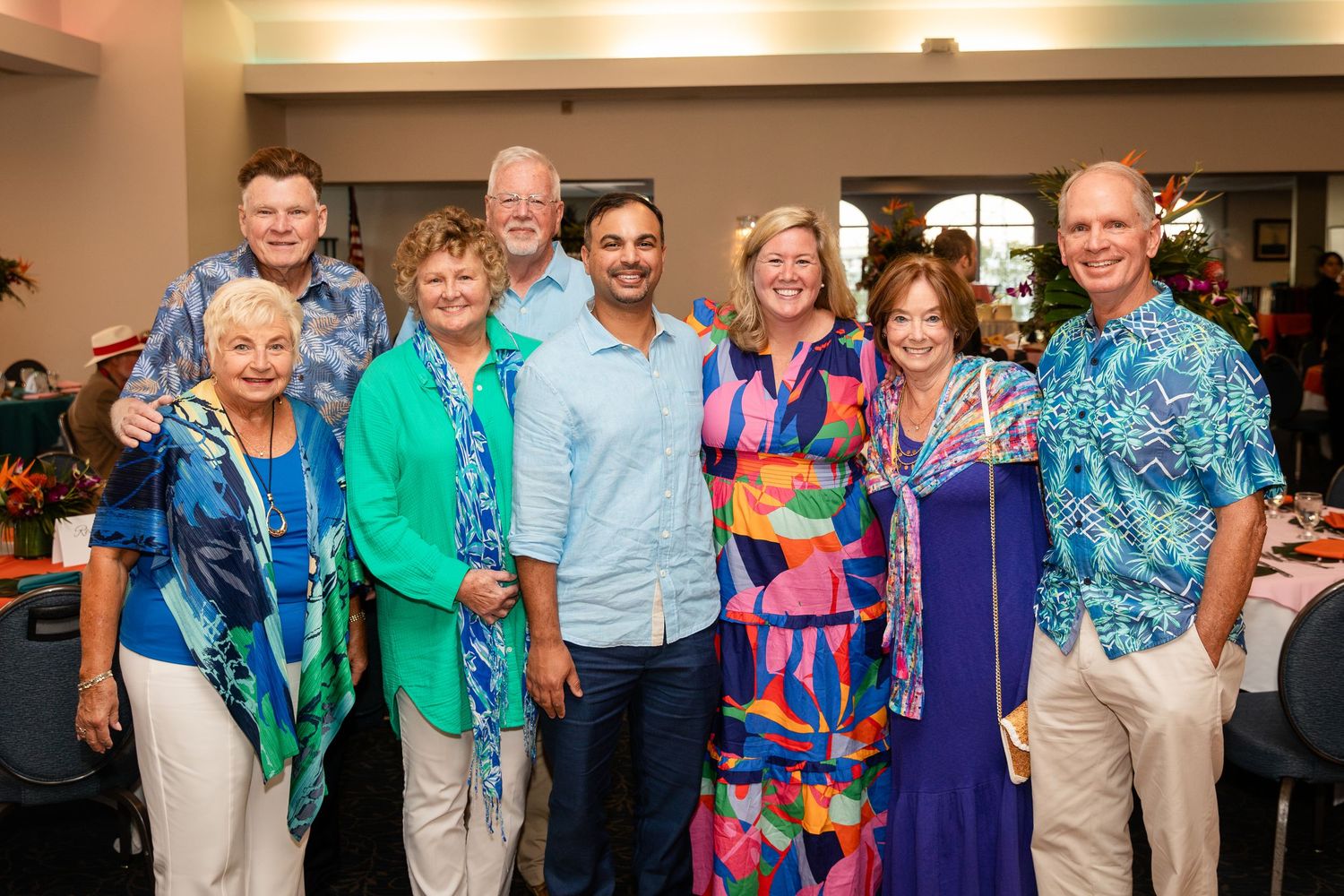 A cheerful group of eight adults posing together at a colorful tropical-themed indoor social gathering event.