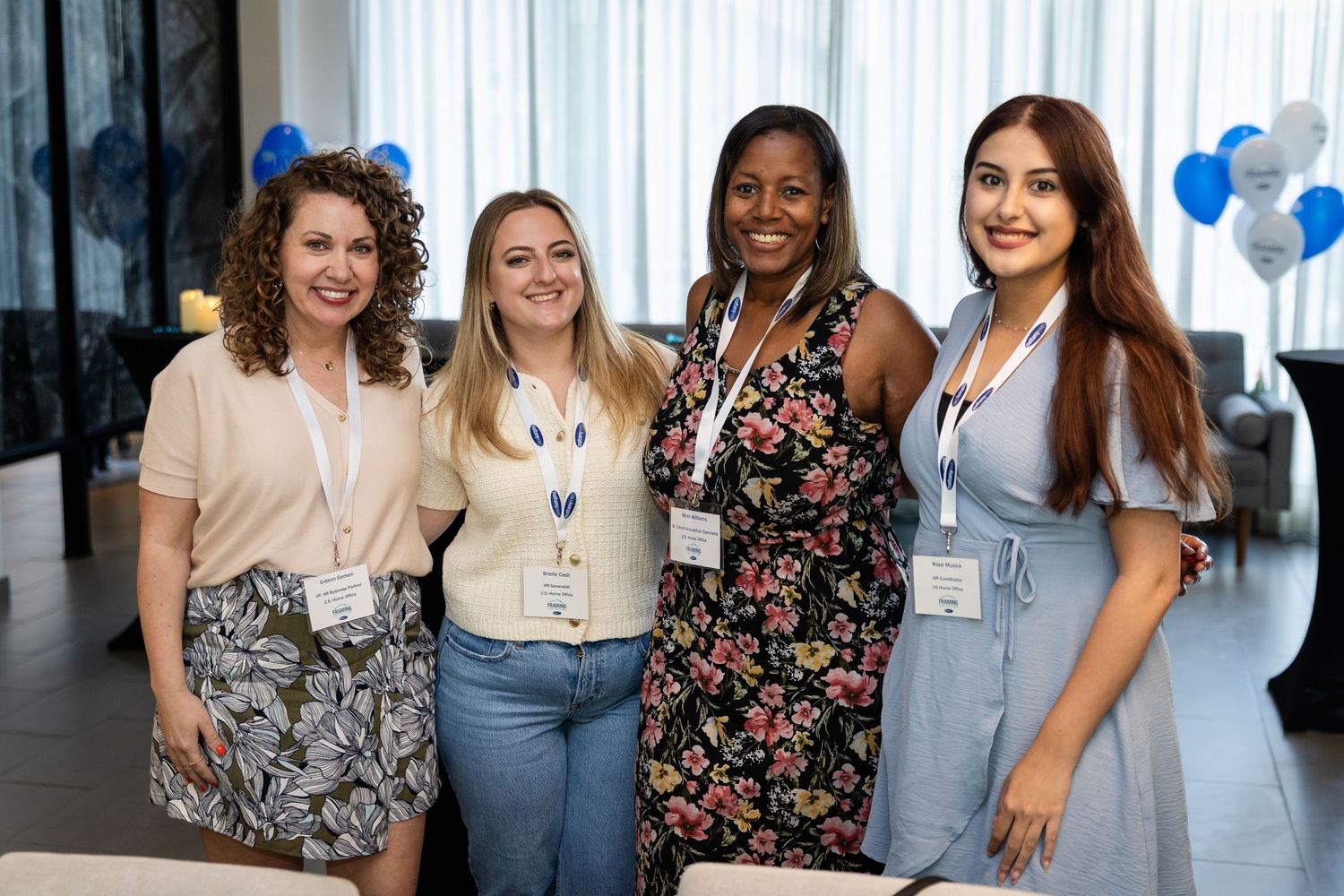Four women smiling together at a corporate event with blue and white balloon decorations in the background.