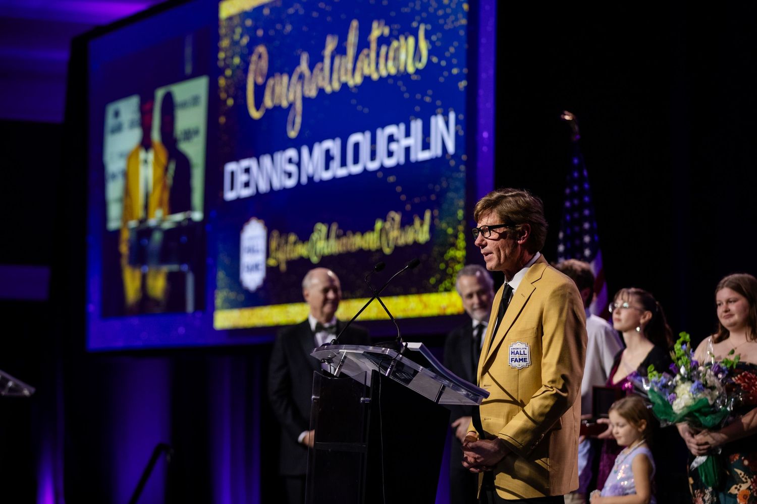 Dennis McLoughlin receives Lifetime Achievement Award on stage, wearing gold blazer at podium with congratulations screen.