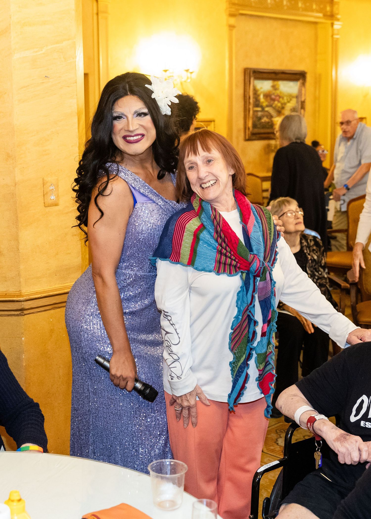 Drag queen in purple gown poses warmly with smiling guest at an indoor Pride event.