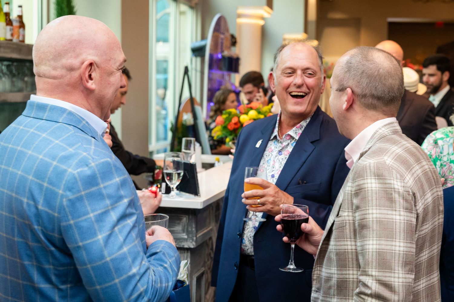 Three men in suits laughing and socializing at a formal networking event, holding wine glasses near a bar.