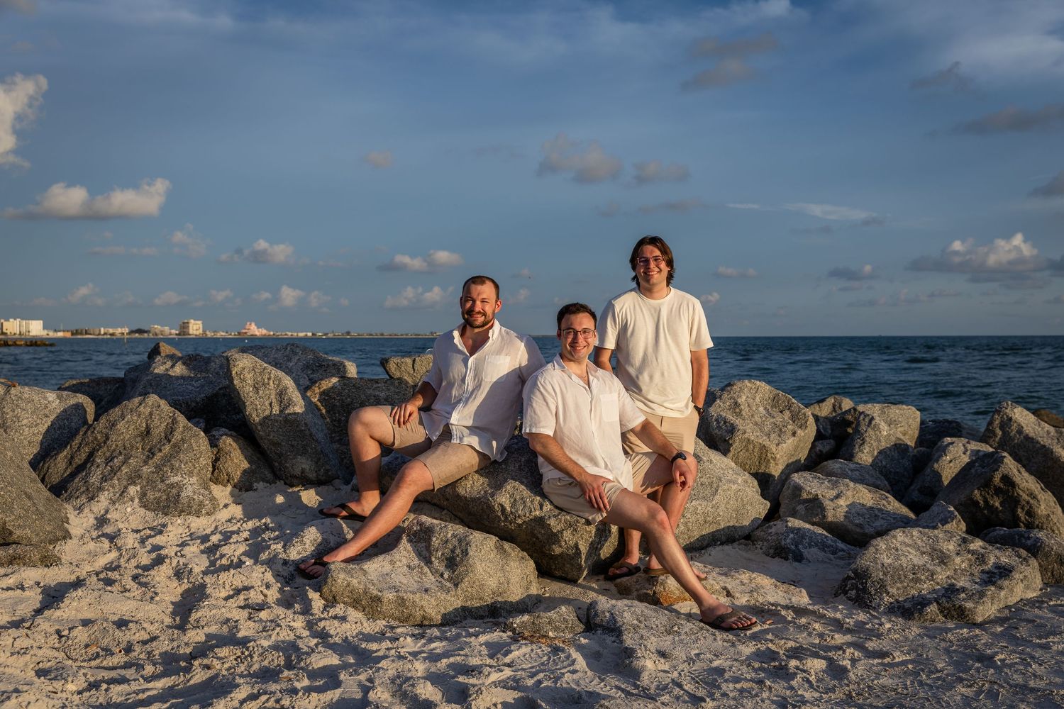 Three men posing on rocky shoreline at sunset with ocean and blue sky in background.