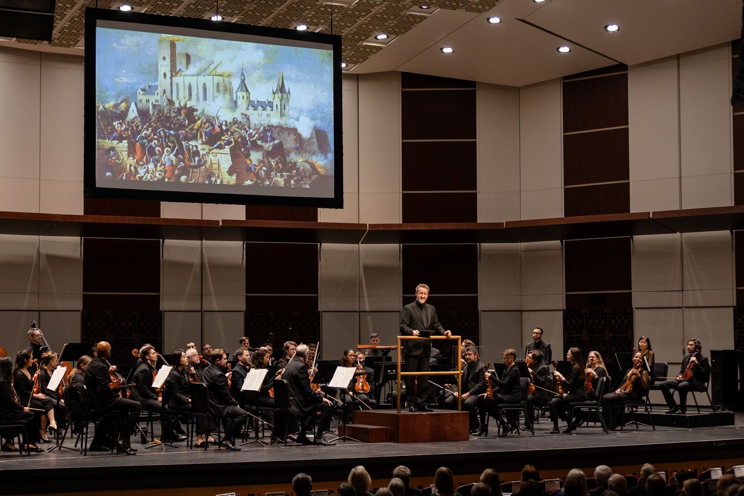 Orchestra performs on stage beneath large projected painting in a concert hall with audience.