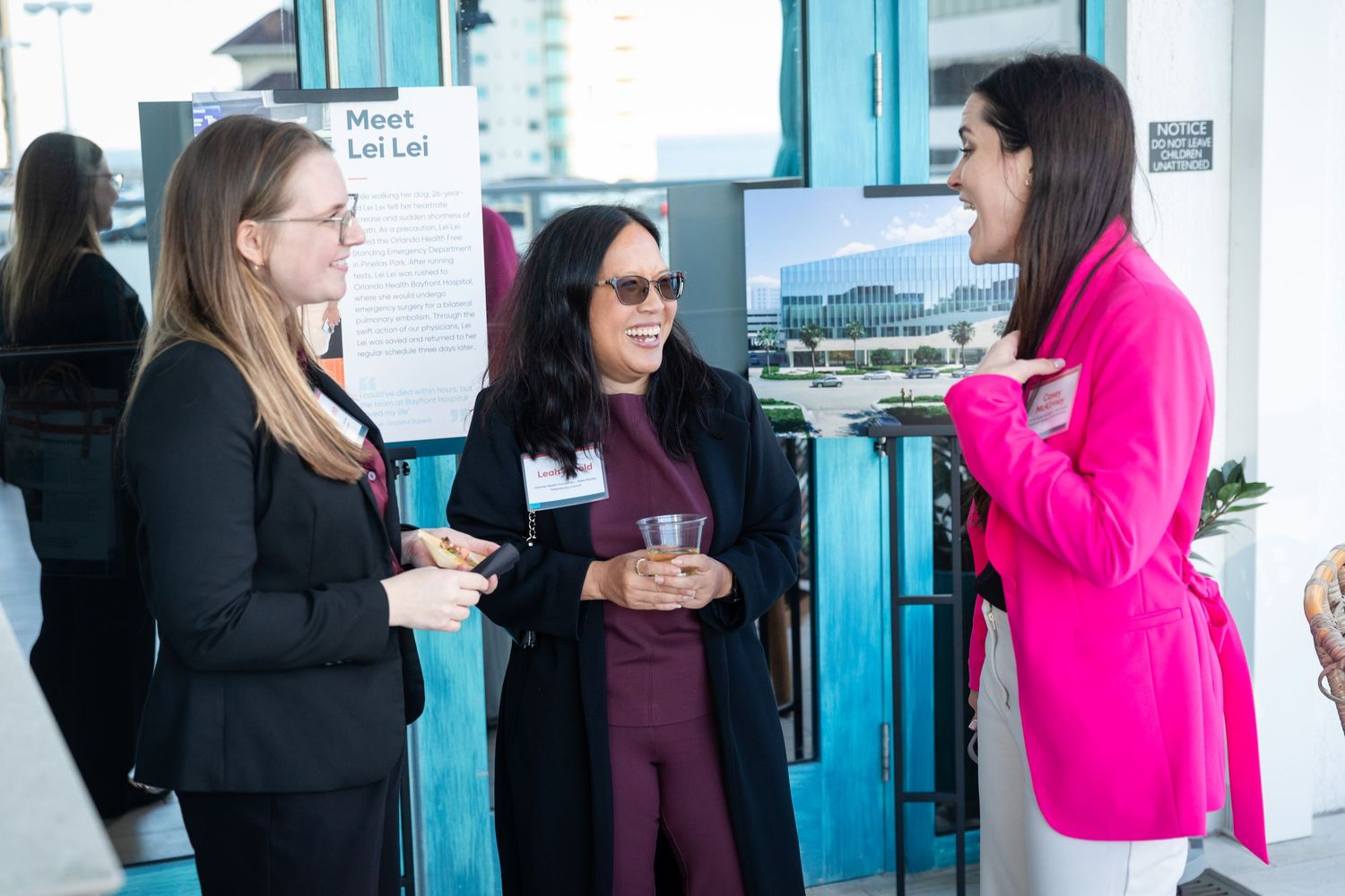 Three women networking at a professional event near a Meet Lei Lei display board and architectural photos.