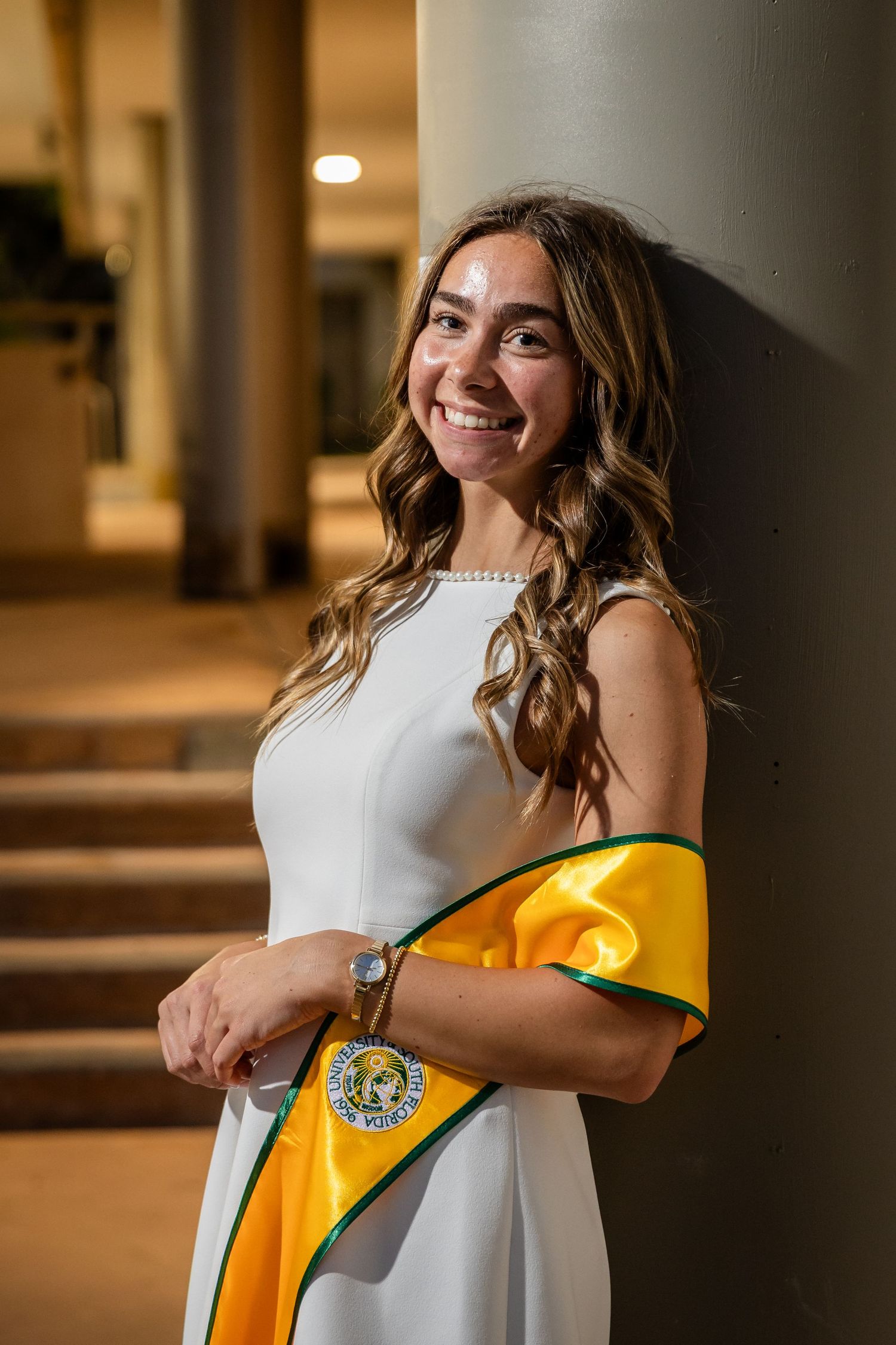 Smiling young woman in white dress with gold and green graduation sash leaning against a pillar.