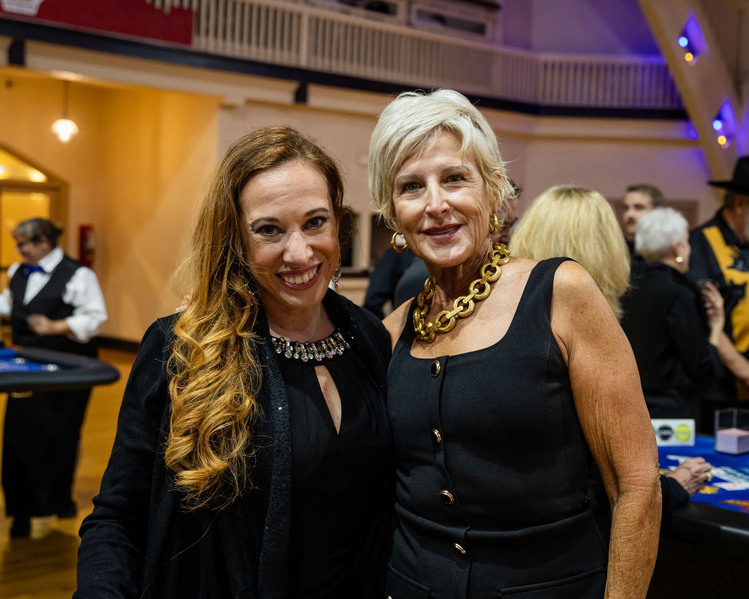 Two women in black formal attire smile together at an elegant casino charity fundraiser event.