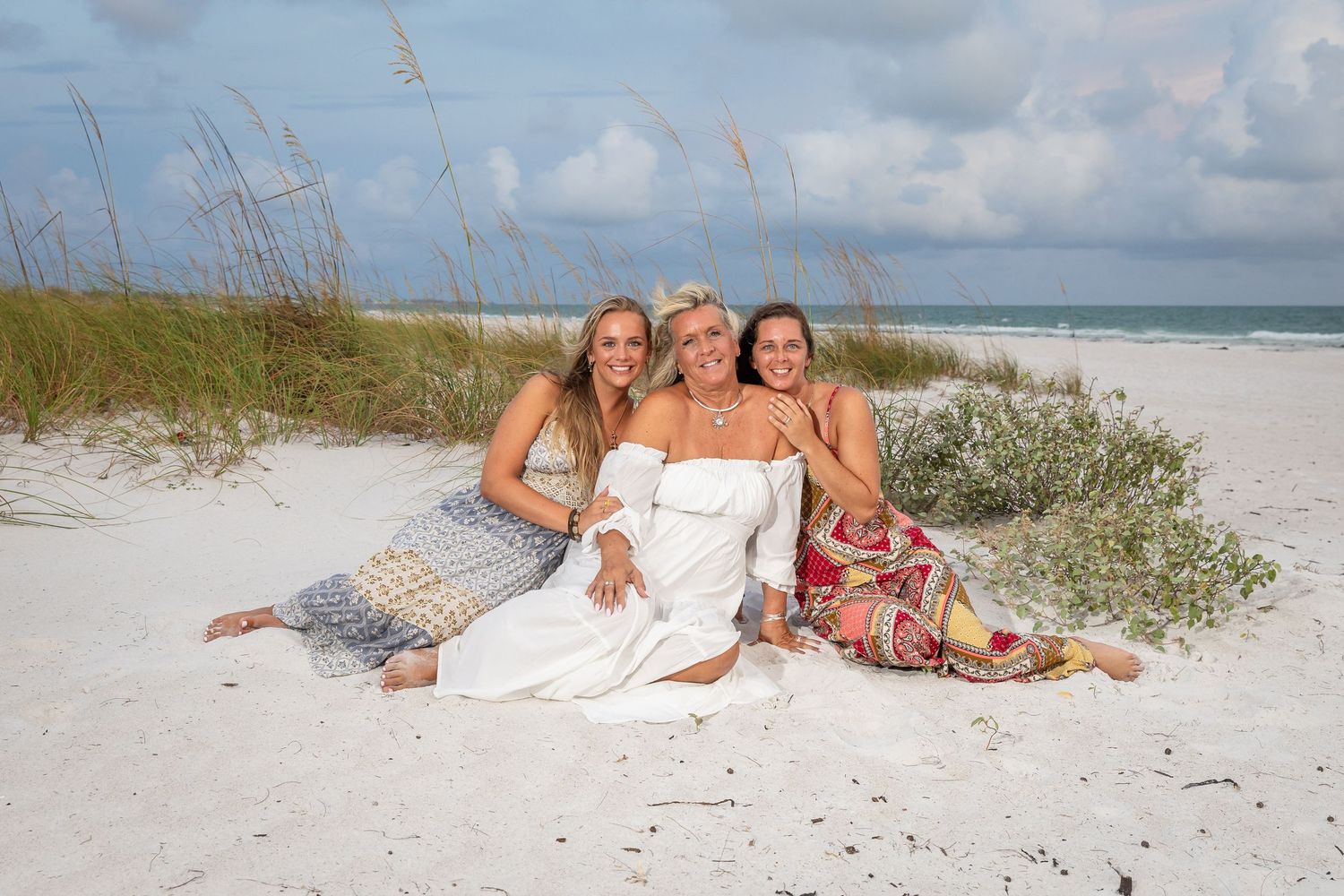 Three women smiling and posing together on white sandy beach near sea oats and sand dunes.