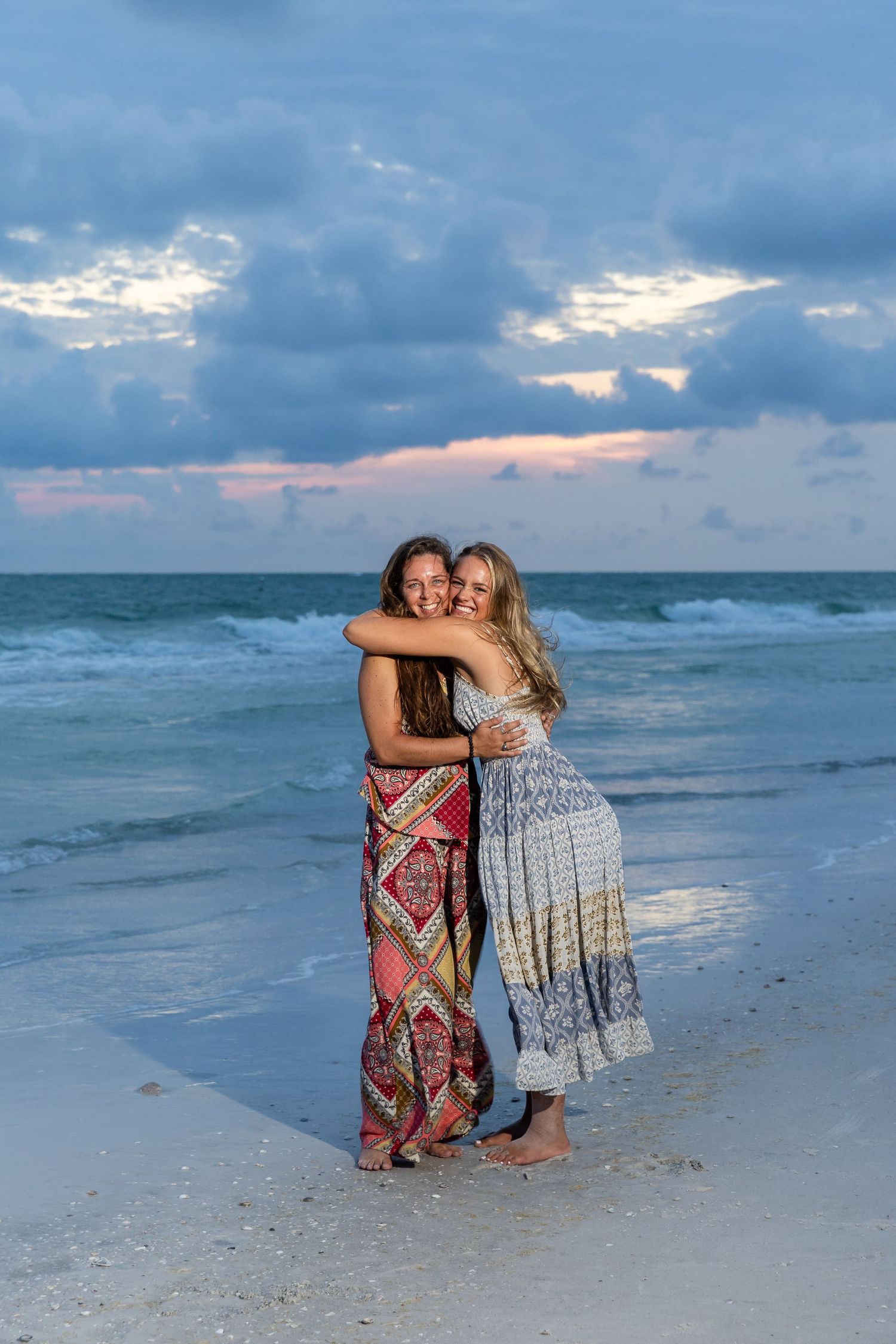 Two women hugging on a sandy beach at sunset, wearing colorful maxi dresses with ocean waves behind them.