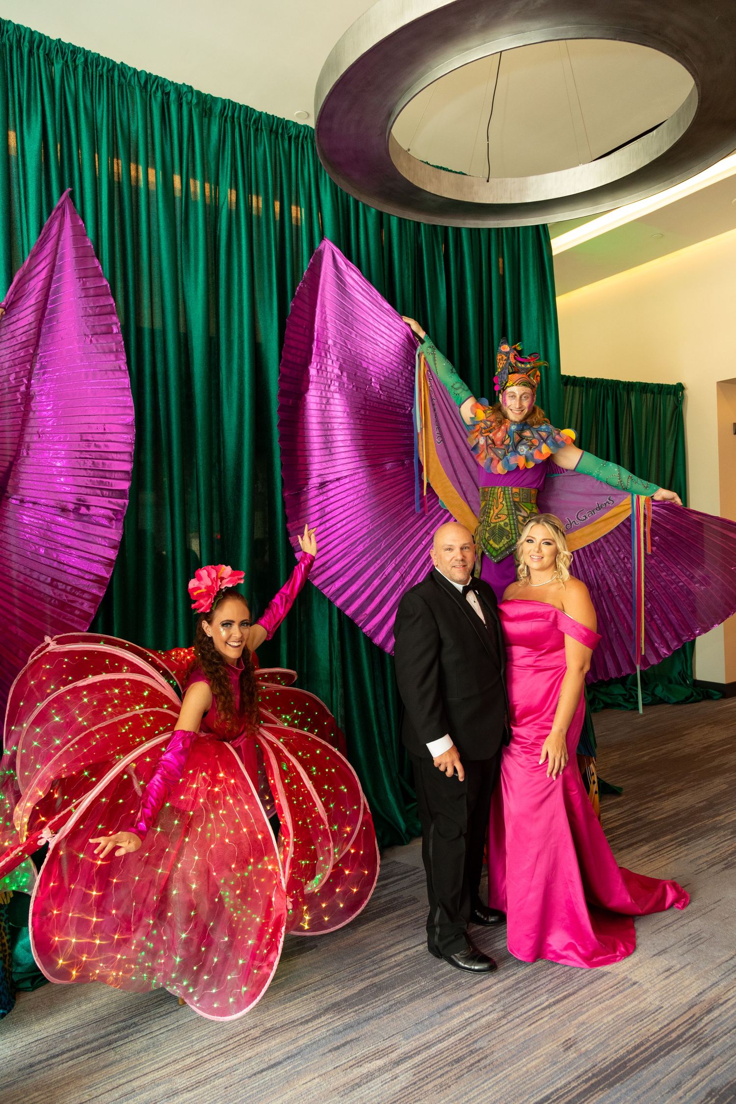 Performers in vibrant costumes pose with elegantly dressed couple at colorful gala event with green drape backdrop.