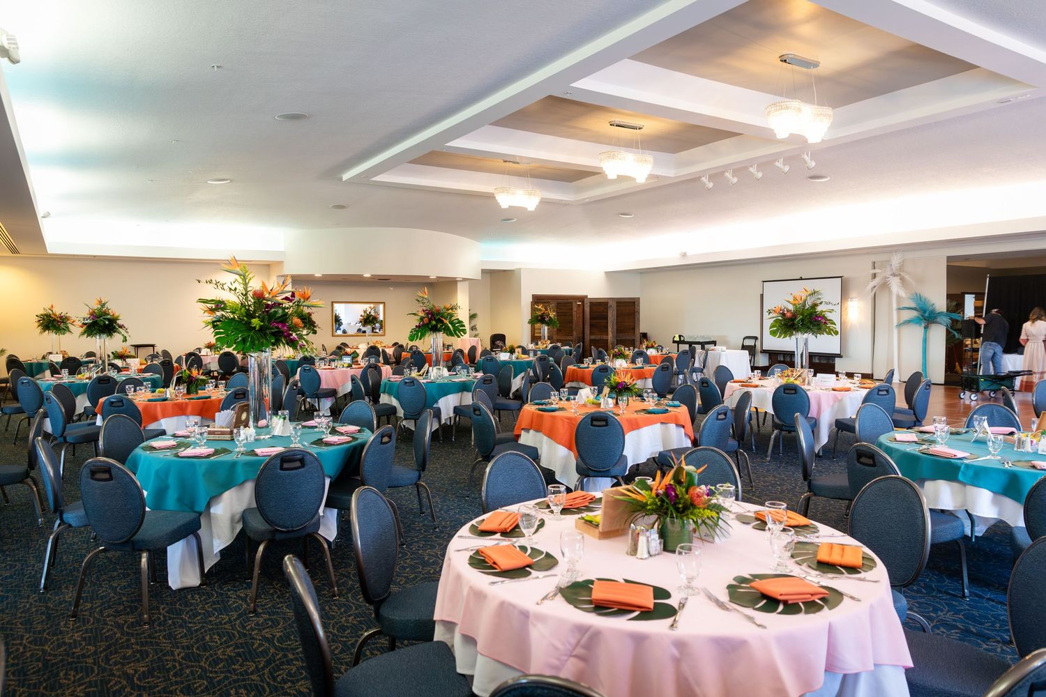 Elegant banquet hall with round tables set for a event, featuring teal and orange linens, floral centerpieces, and gray chairs.