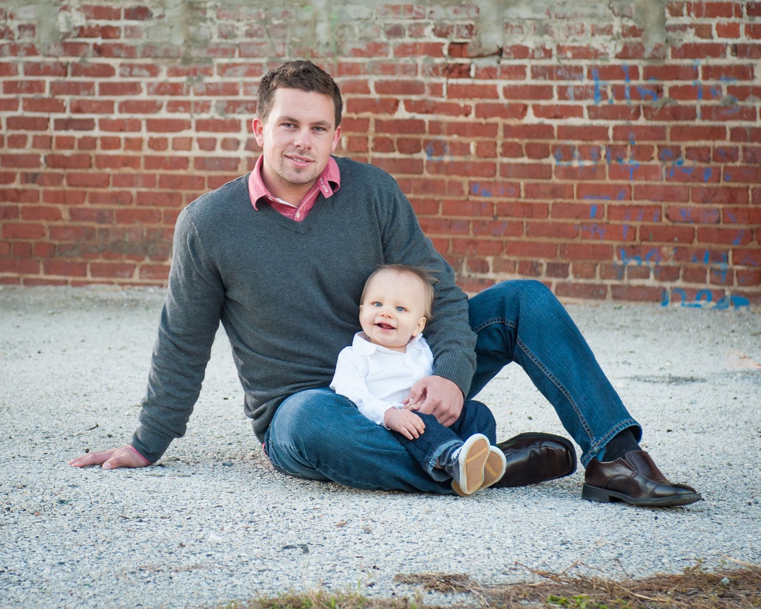 A young man sits on gravel against a brick wall, smiling and holding a happy baby in a white outfit.