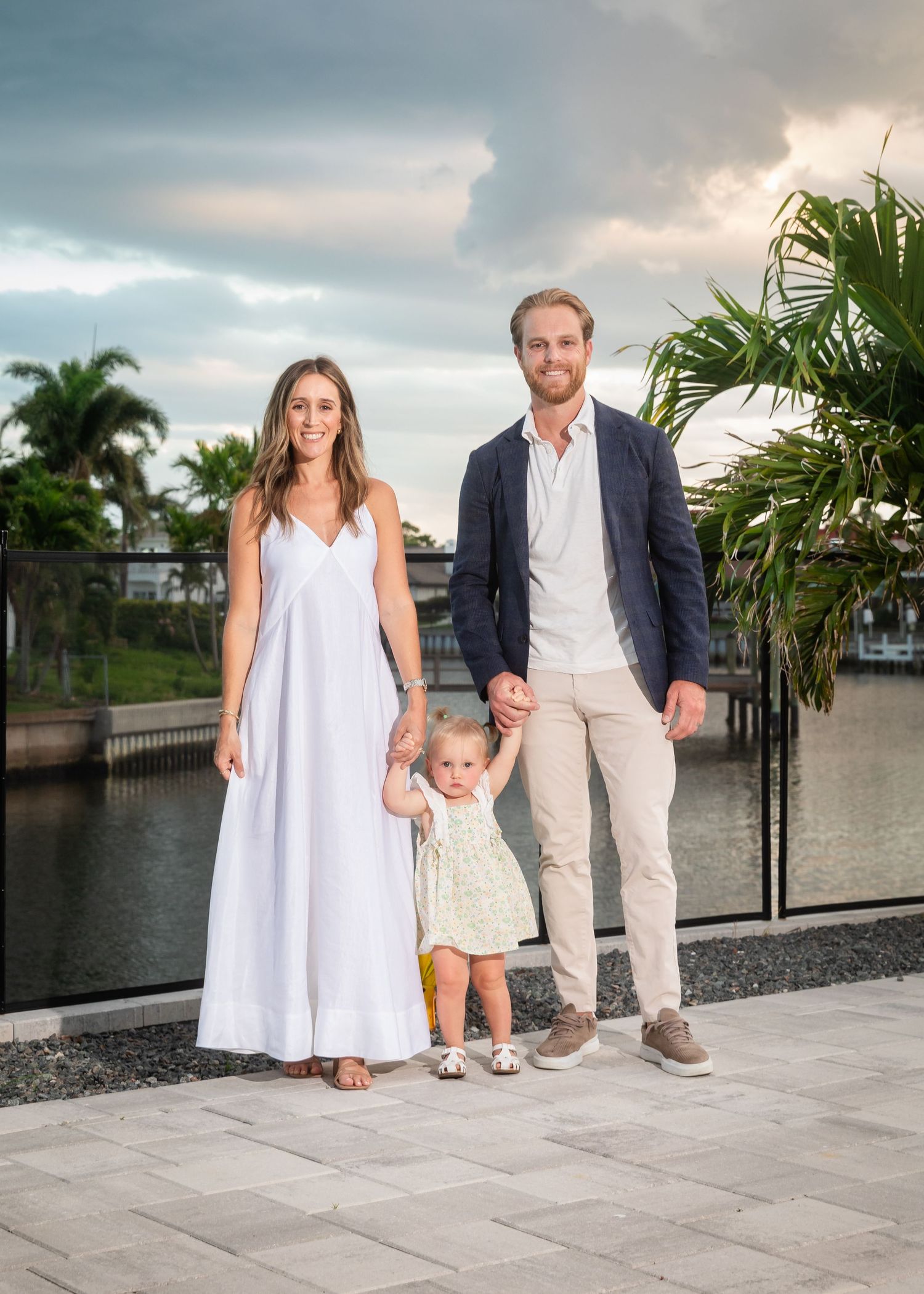 A family of three poses outdoors near a waterway with palm trees, parents holding a toddler girl between them.