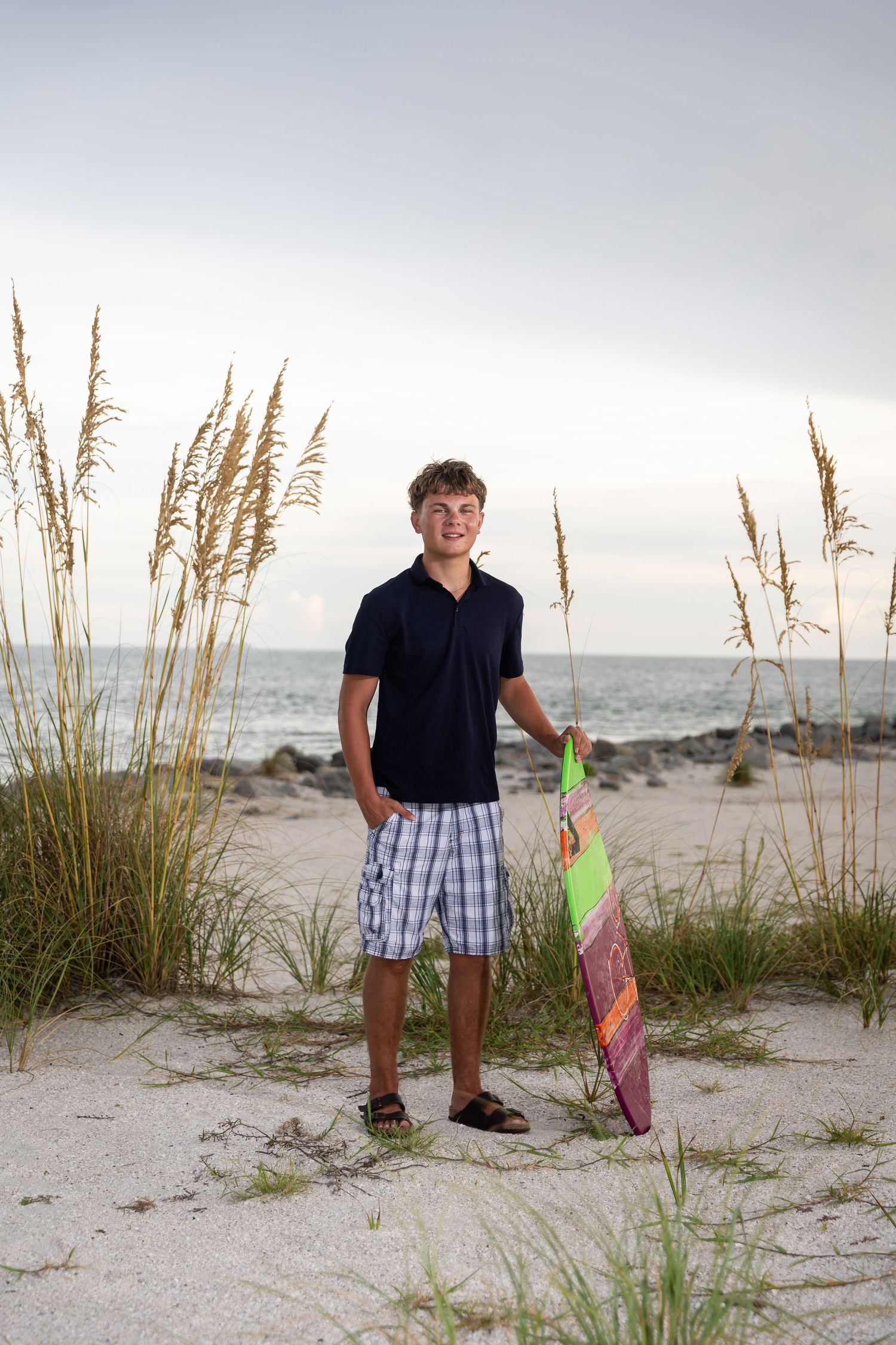 Teen boy holding a colorful skateboard on a sandy beach with sea oats, wearing plaid shorts and a navy polo shirt.