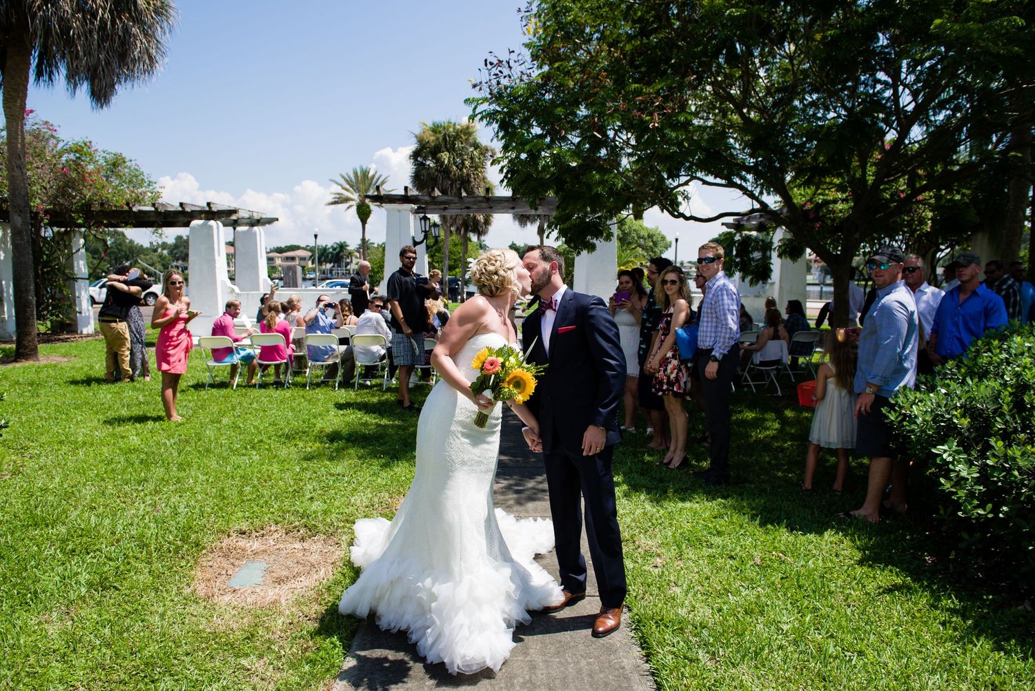 Bride and groom share a kiss at an outdoor wedding ceremony on a sunny day with guests seated on chairs.