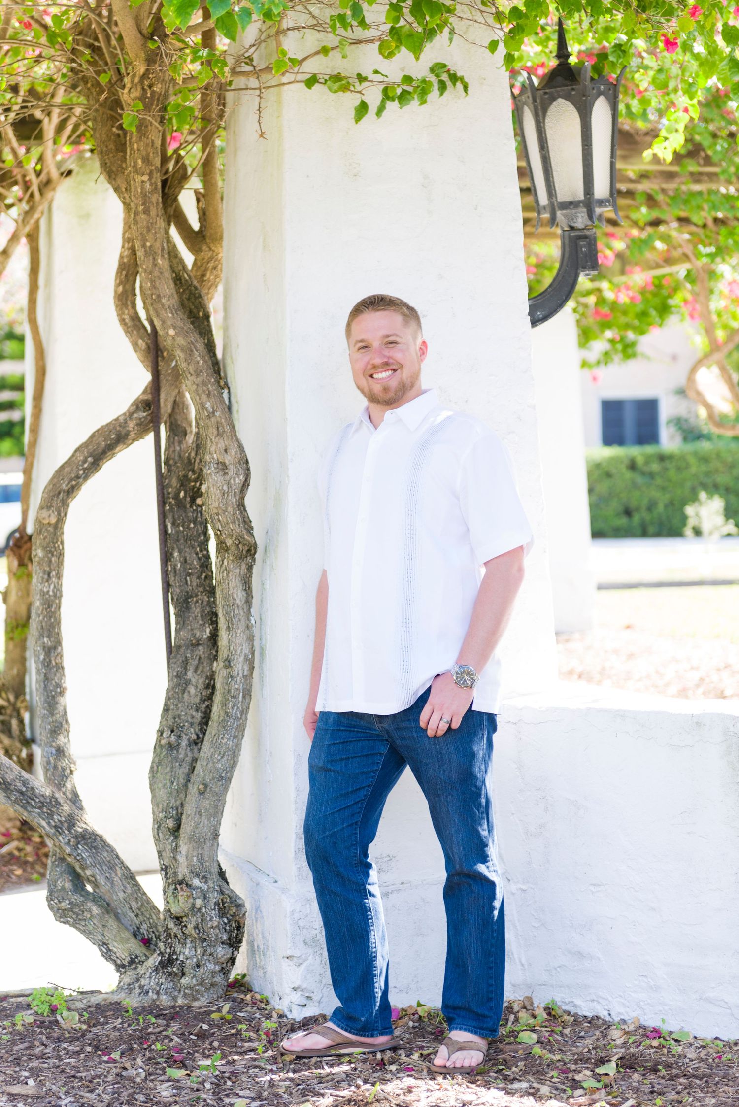 A smiling man in a white shirt and jeans leans against a white pillar surrounded by lush greenery.