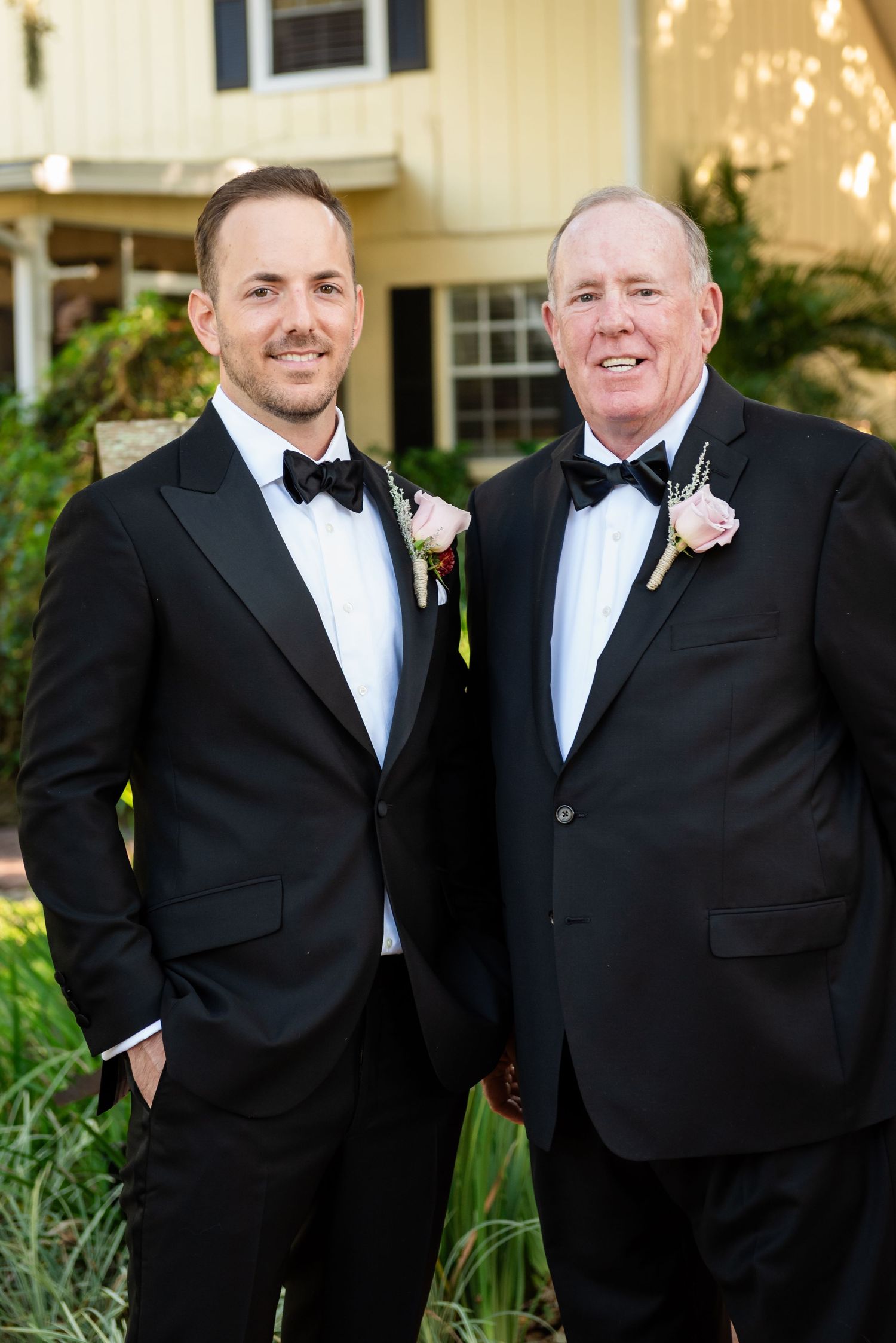 Two men in matching black tuxedos with boutonnieres pose together outdoors at a wedding celebration.