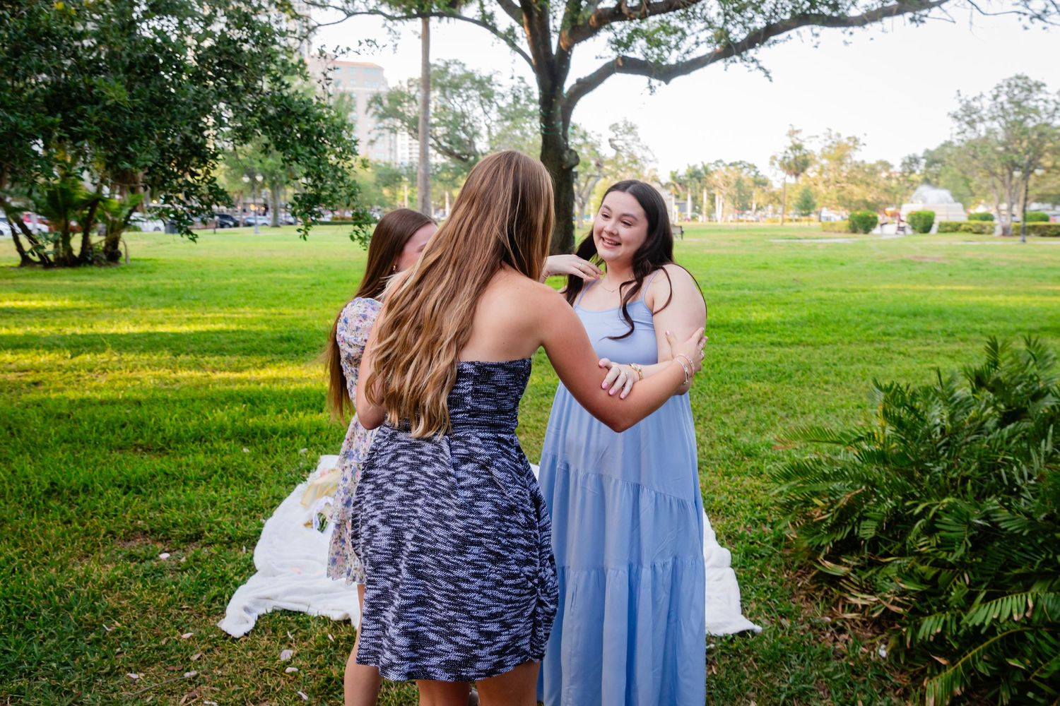 Three young women laughing and embracing together in a sunny green park during a casual outdoor gathering.