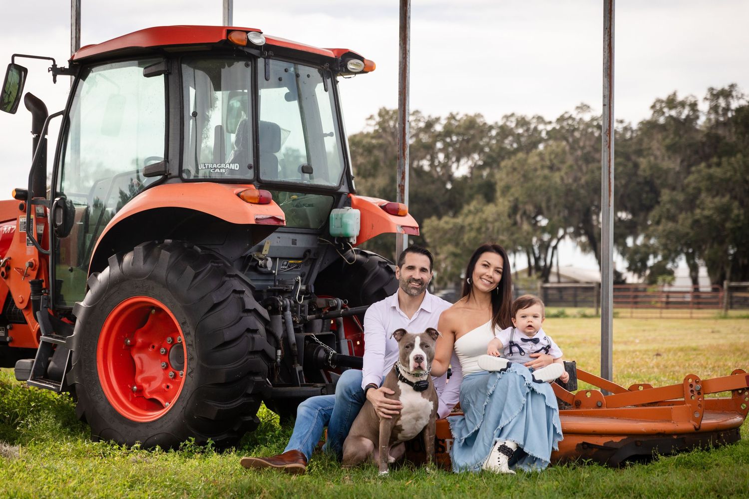 Family with dog poses in front of orange Kubota tractor on green grass farm field for portrait photo.