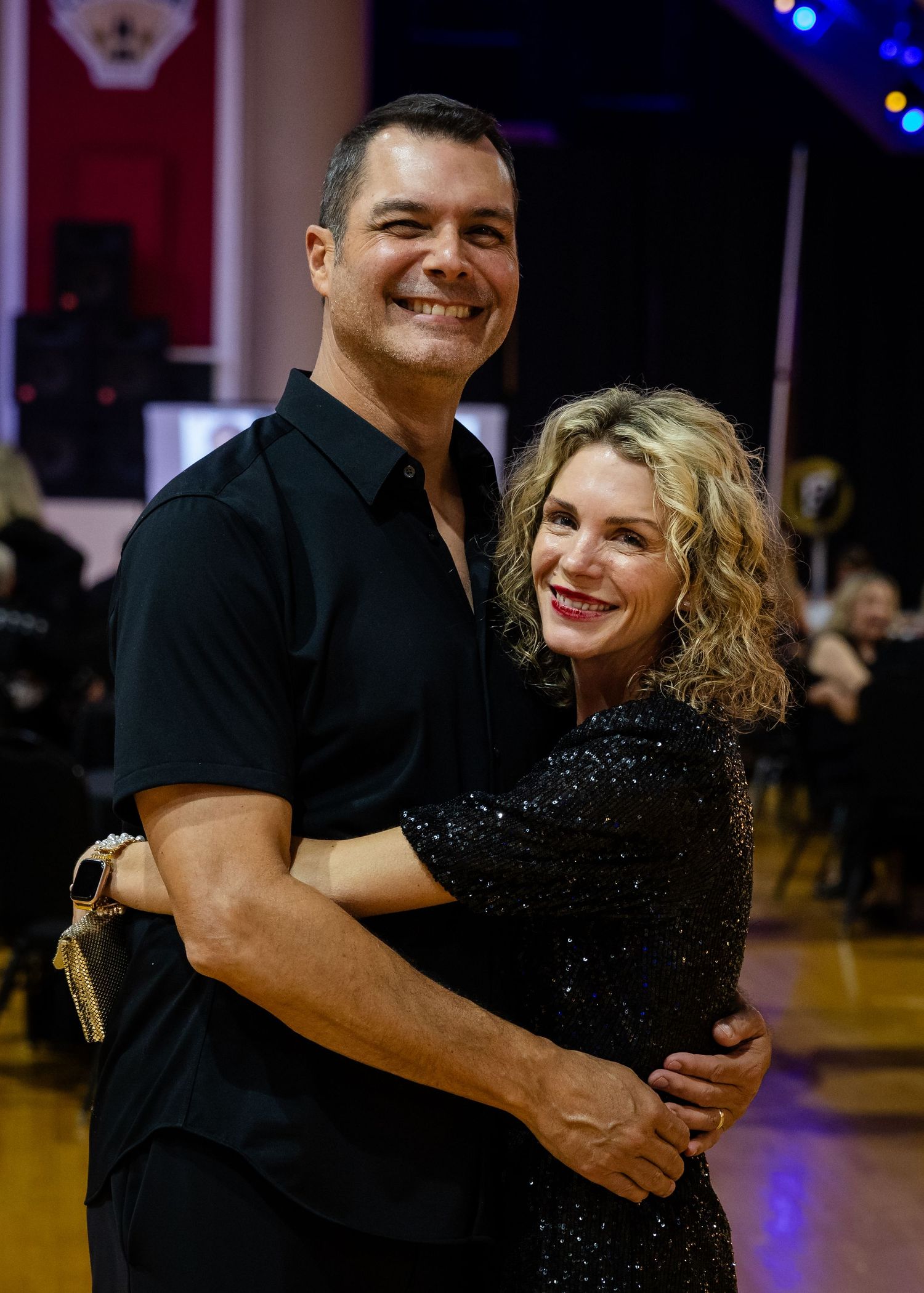 Smiling couple dressed in black embraces on a ballroom dance floor at an elegant evening event.