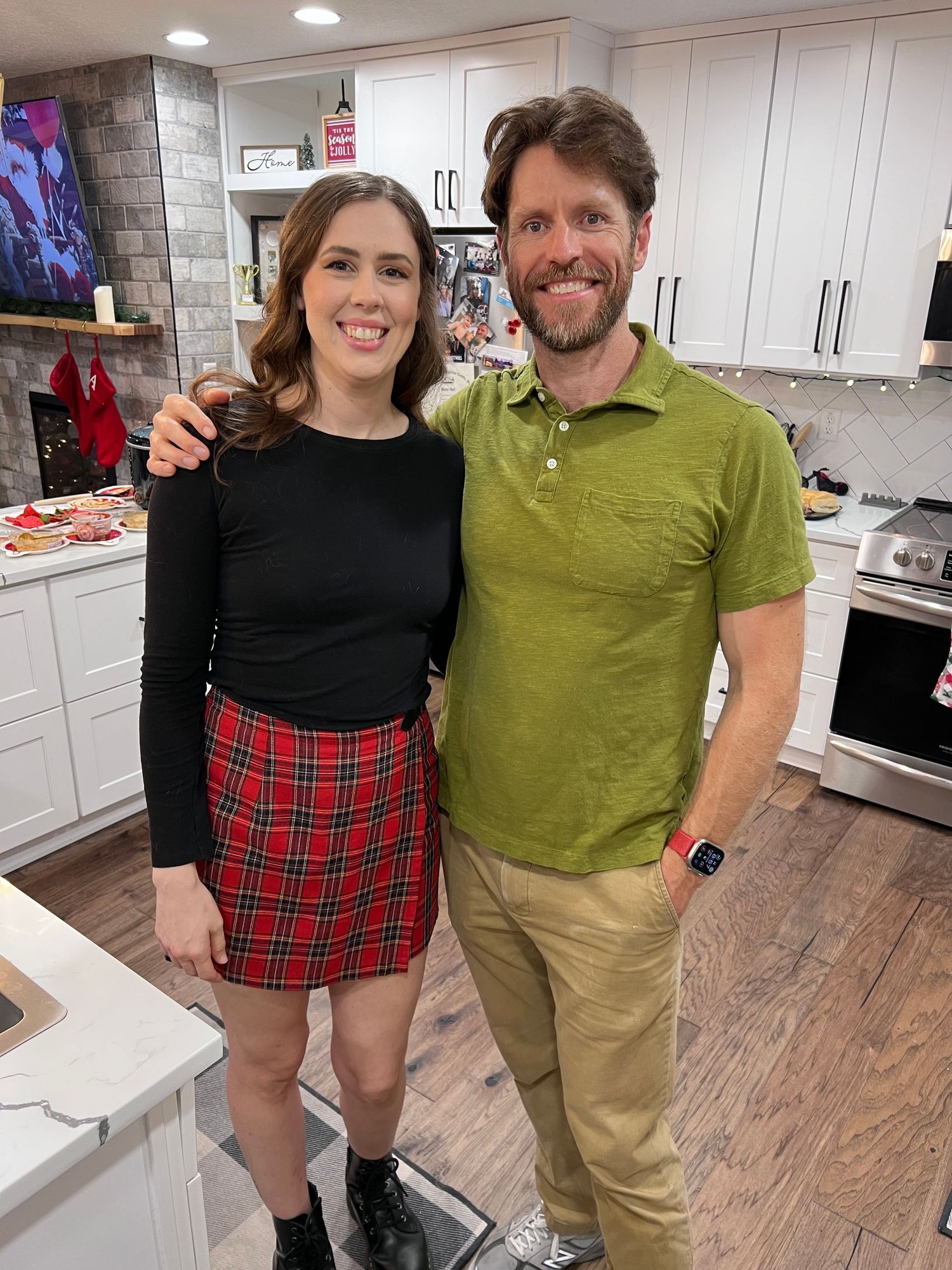 A smiling couple poses together in a modern kitchen, woman in black top and plaid skirt, man in green polo shirt.