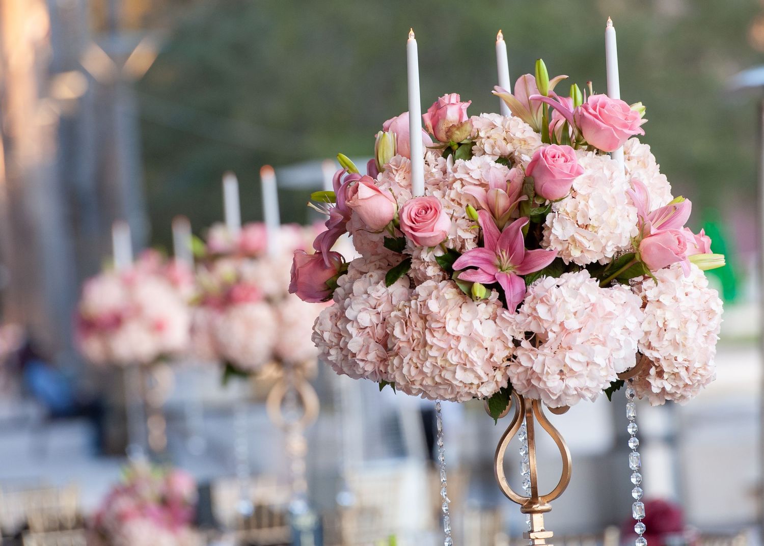 Elegant wedding centerpiece with pink roses, hydrangeas, lilies, and white taper candles on a gold stand.