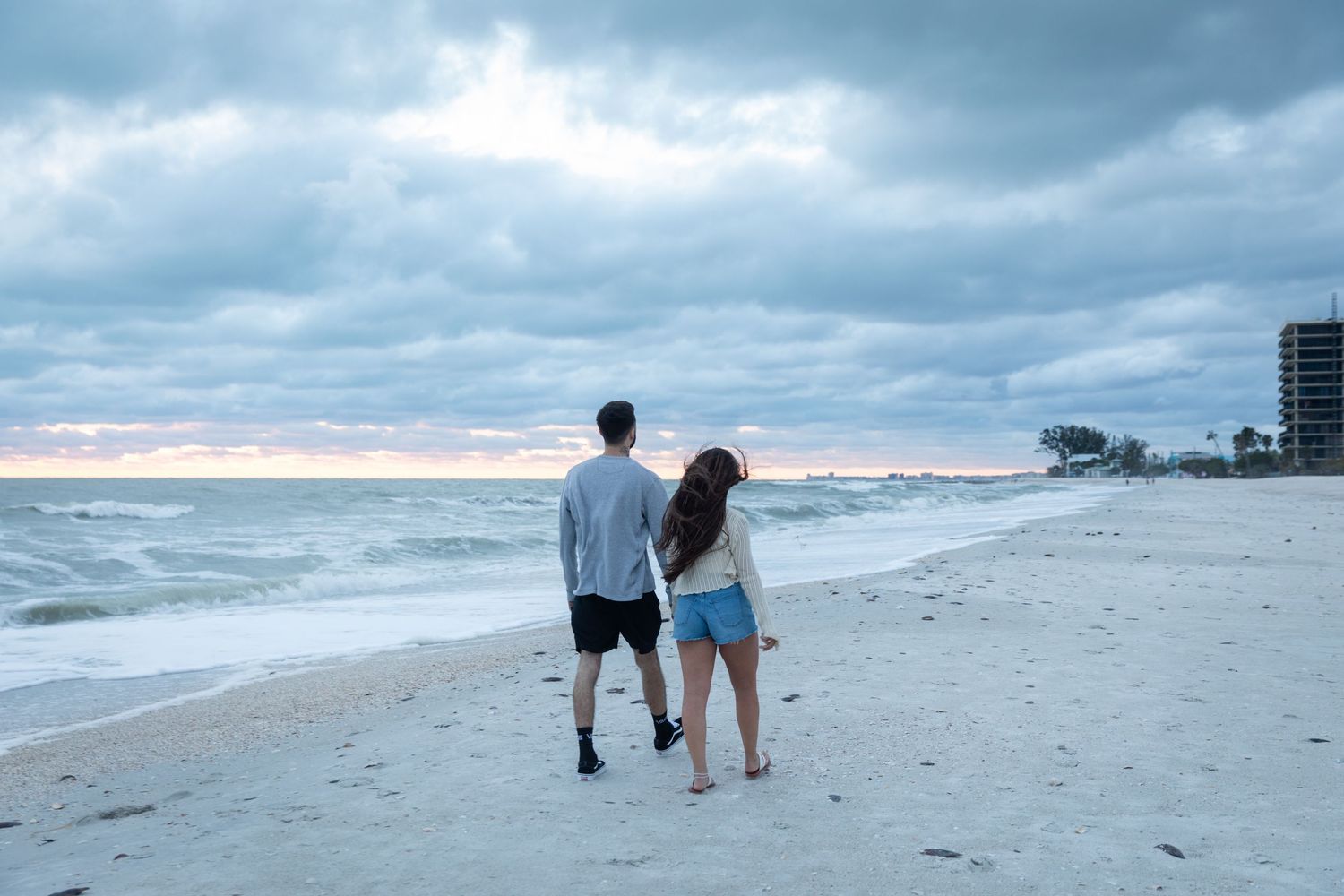 Couple walking hand in hand along a sandy beach at dusk with cloudy skies overhead.