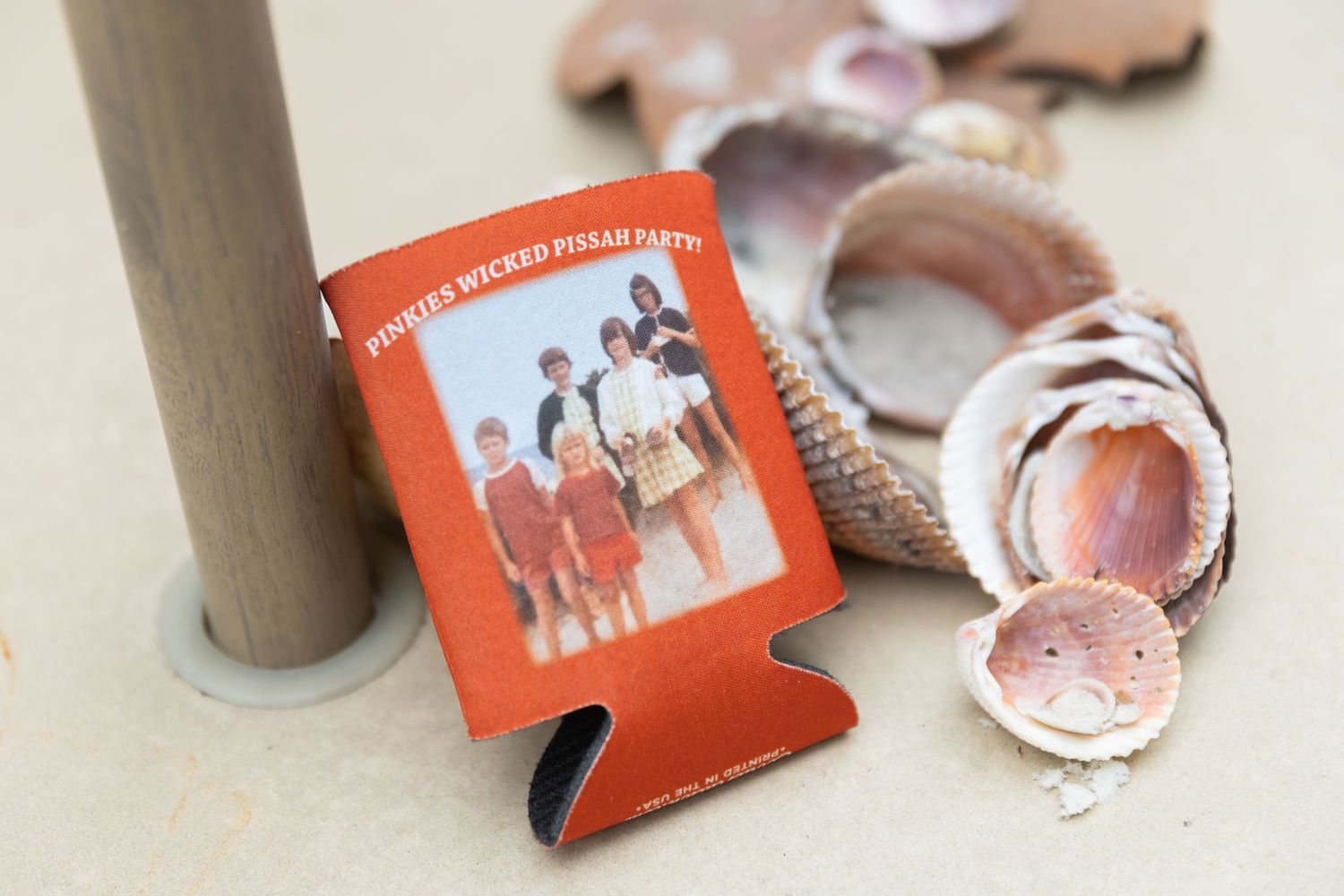 Orange koozie with retro photo and text near seashells on sandy surface.