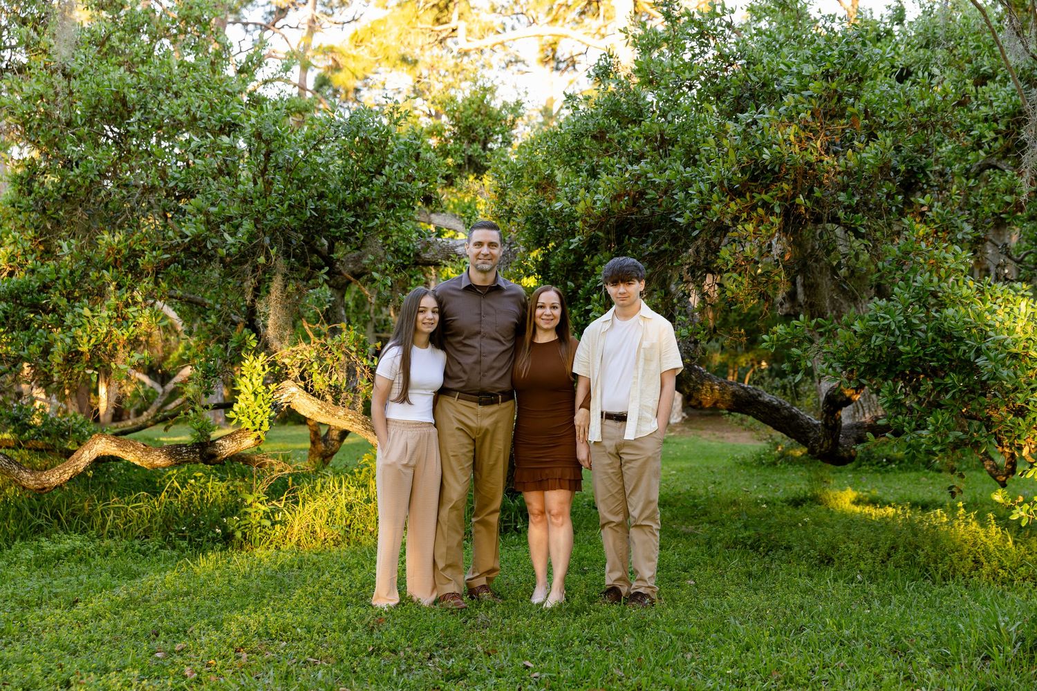 Family of four smiling together outdoors in a lush green garden with trees in warm golden light.